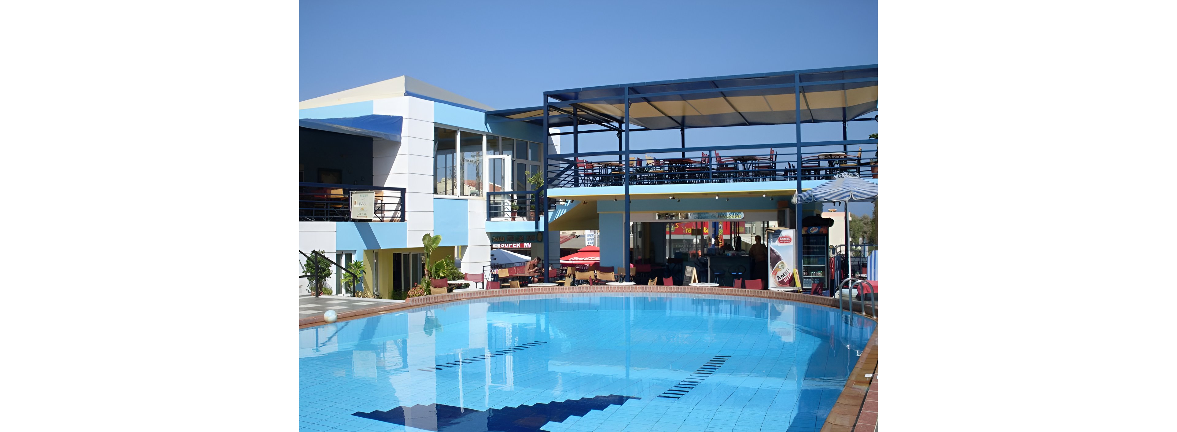 A clear blue swimming pool sits in front of a modern hotel or resort building. The structure features multiple levels with balconies, large windows, and an elevated open-air bar or dining area. Bright sun and a cloudless sky create a warm, inviting atmosphere. Pool ladders, tiled edges, and surrounding plants emphasize a relaxed vacation setting.
