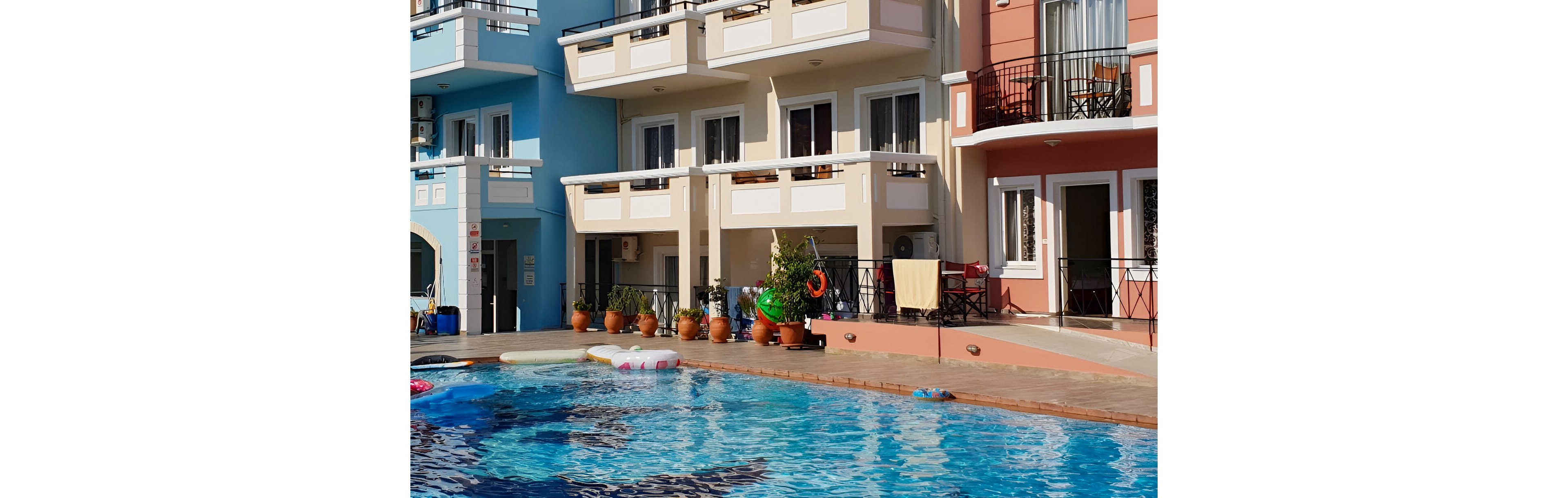 A pastel-colored hotel building features multiple balconies overlooking a sunlit outdoor swimming pool. Several people, including children and adults, are enjoying the water and relaxing poolside. Potted plants and pool floats add to the lively, vacation atmosphere. The architecture includes arched windows and geometric shapes, with clear blue skies above. No visible text or numbers are present in the image.
