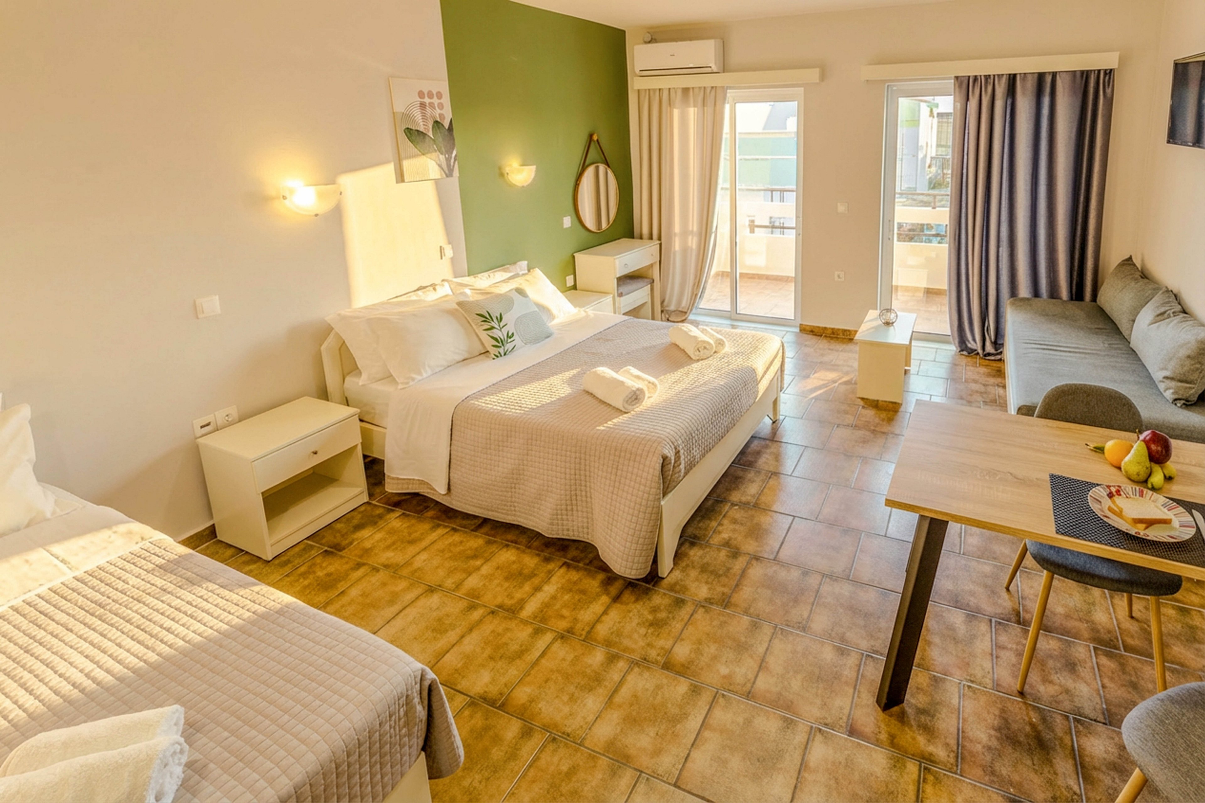 Spacious hotel room featuring a double bed and a single bed, both neatly made with neutral bedding. The room is well-lit with natural light from large sliding glass doors leading to a balcony. Key visuals include a small dining table with fruit, a sofa, and a green accent wall. The decor is minimalistic with warm tones and contemporary furnishings.
