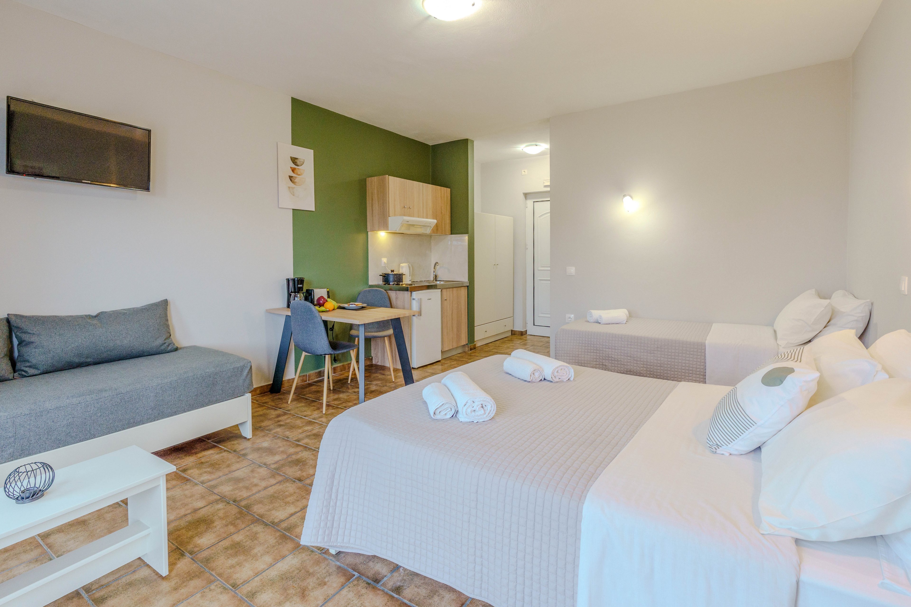 A spacious studio apartment features two beds and a sofa, with a small kitchenette and dining area in the background. The room is decorated in neutral tones with green accent walls and tiled flooring. Towels are neatly arranged on the beds, and a flat-screen TV is mounted on the wall. The space is well-lit, creating a welcoming and comfortable atmosphere.