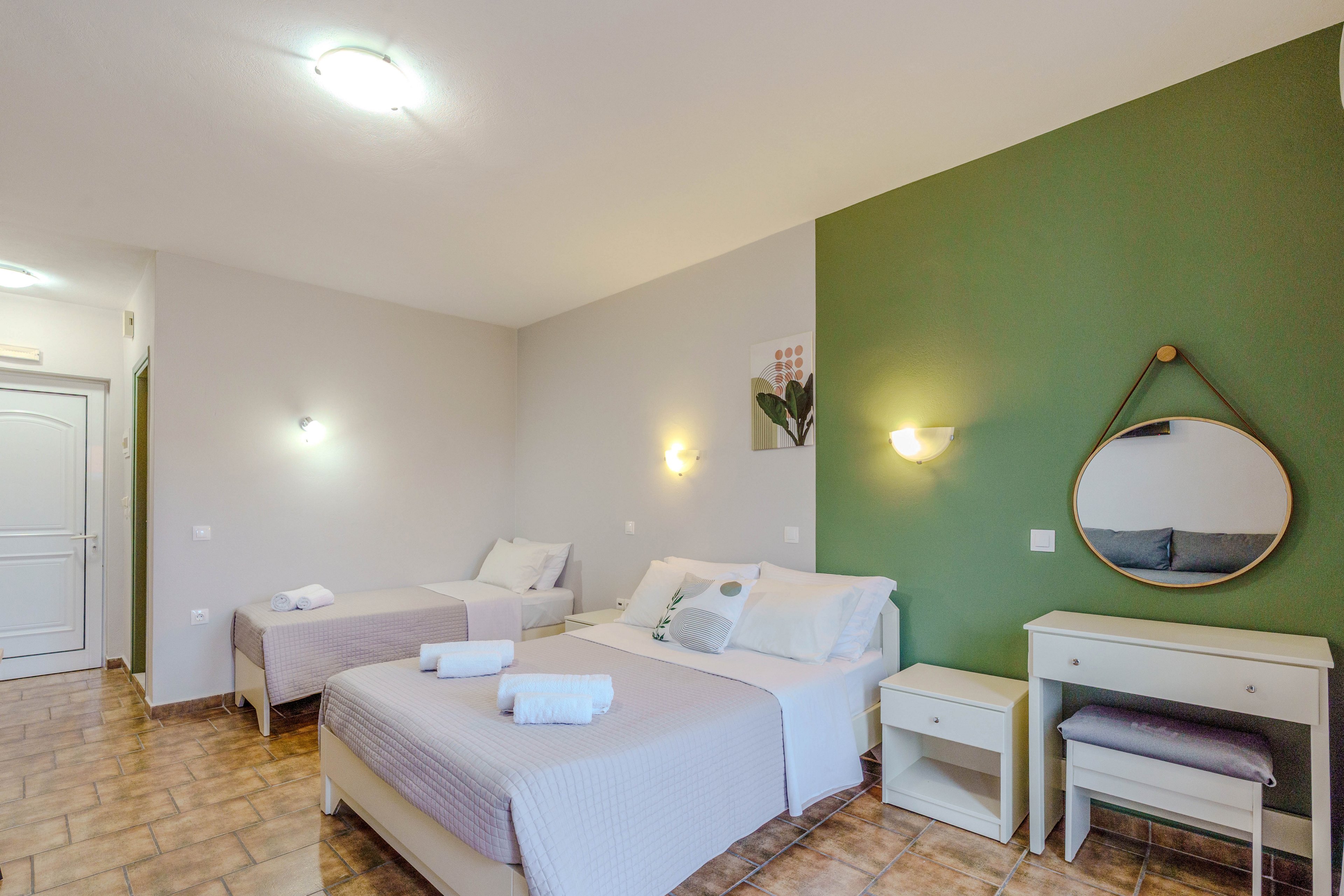 A spacious, well-lit hotel room features a double bed and a single bed, both neatly made with white linens and towels. The room has a green accent wall with a round mirror above a small vanity desk and stool. Warm lighting and minimalist decor create a welcoming, contemporary atmosphere. The tiled floor and neutral color palette add to the clean, modern look.