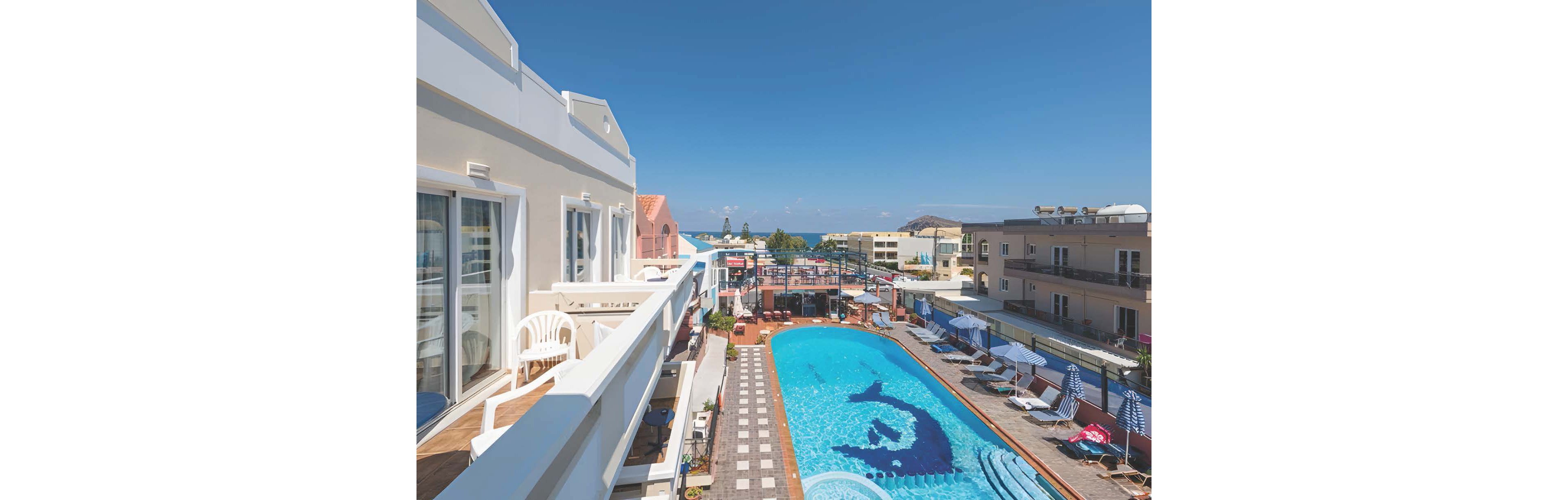 The image shows a vibrant hotel pool area surrounded by sun loungers and umbrellas, with balconies overlooking the scene. The pool features a playful dolphin graphic and is set against a backdrop of clear blue skies and distant ocean views. The setting is bright and inviting, with modern architecture and a relaxed vacation atmosphere. No visible text or numeric values are present in the image.