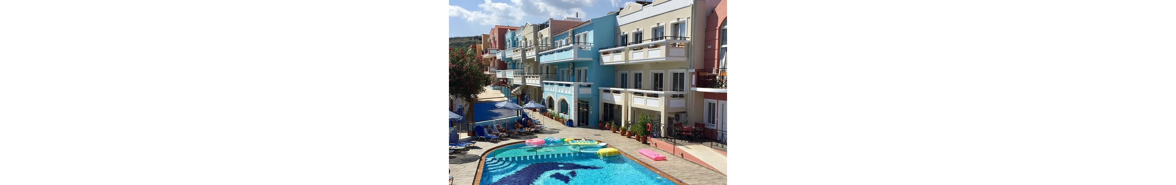 A vibrant outdoor swimming pool is surrounded by pastel-colored hotel buildings under a bright blue sky with scattered clouds. The pool features a large dolphin mosaic and several floating inflatables, with one person swimming. The setting is relaxed and summery, with sun loungers and umbrellas lining the poolside.