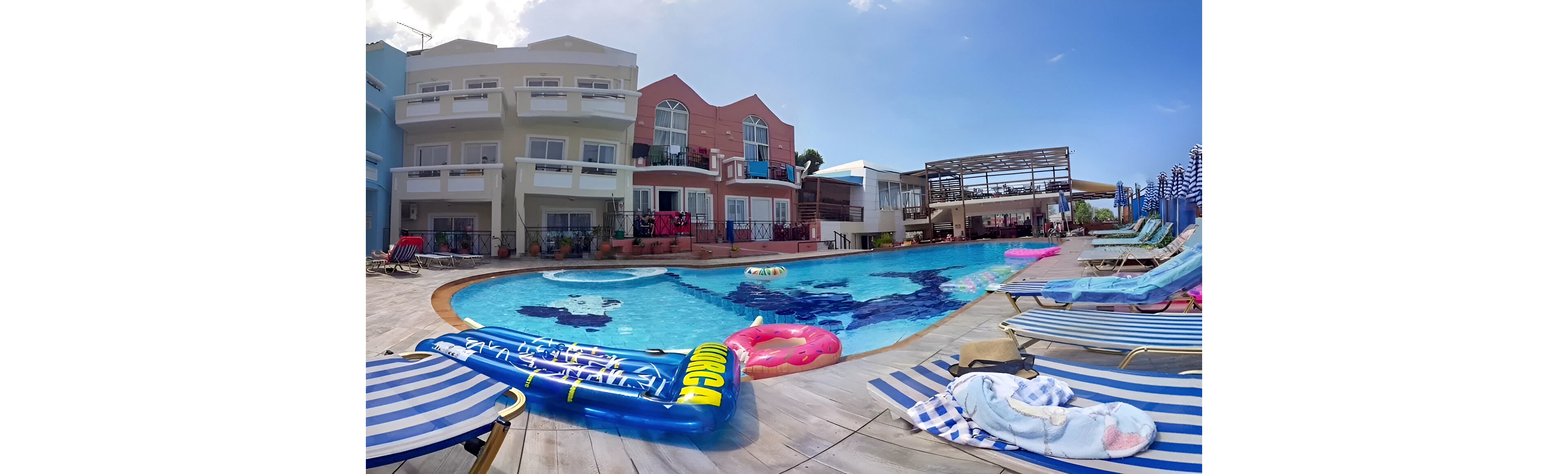 A vibrant outdoor hotel pool is surrounded by striped loungers and colorful inflatable pool floats. Several people relax by the water, enjoying the sunny weather and clear blue sky. The setting features multi-story buildings with balconies and towels, creating a lively vacation atmosphere. The pool area is accented by playful visuals, including a large inflatable with the text 'SUNGA'. The overall mood is relaxed and summery.