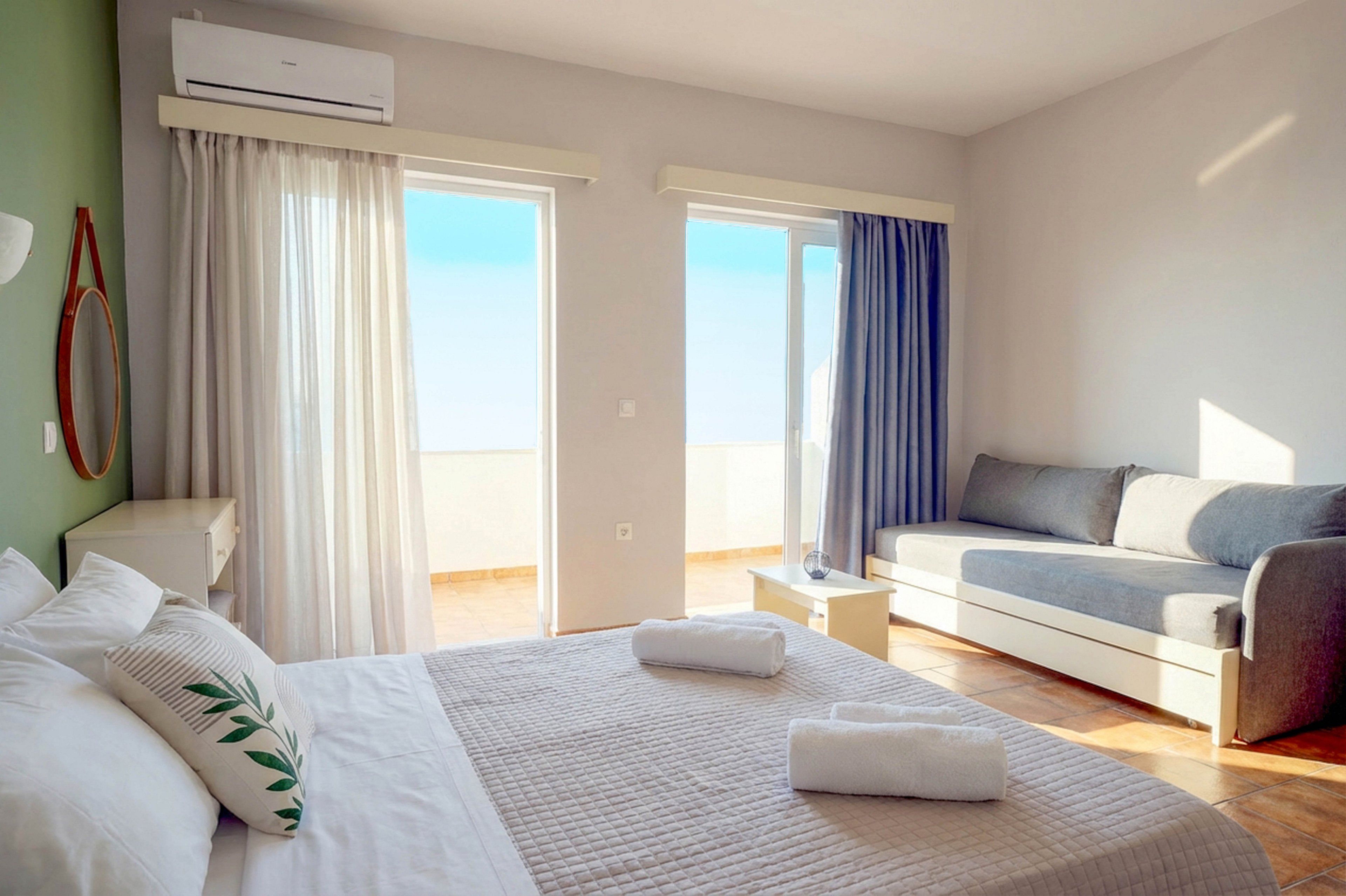 A sunlit hotel room features a neatly made bed with white linens and a decorative pillow, facing a balcony with open glass doors. The space includes a gray sofa, a small table, and an air conditioning unit mounted above the window. Soft natural light fills the room, highlighting the minimalist decor and wooden flooring.