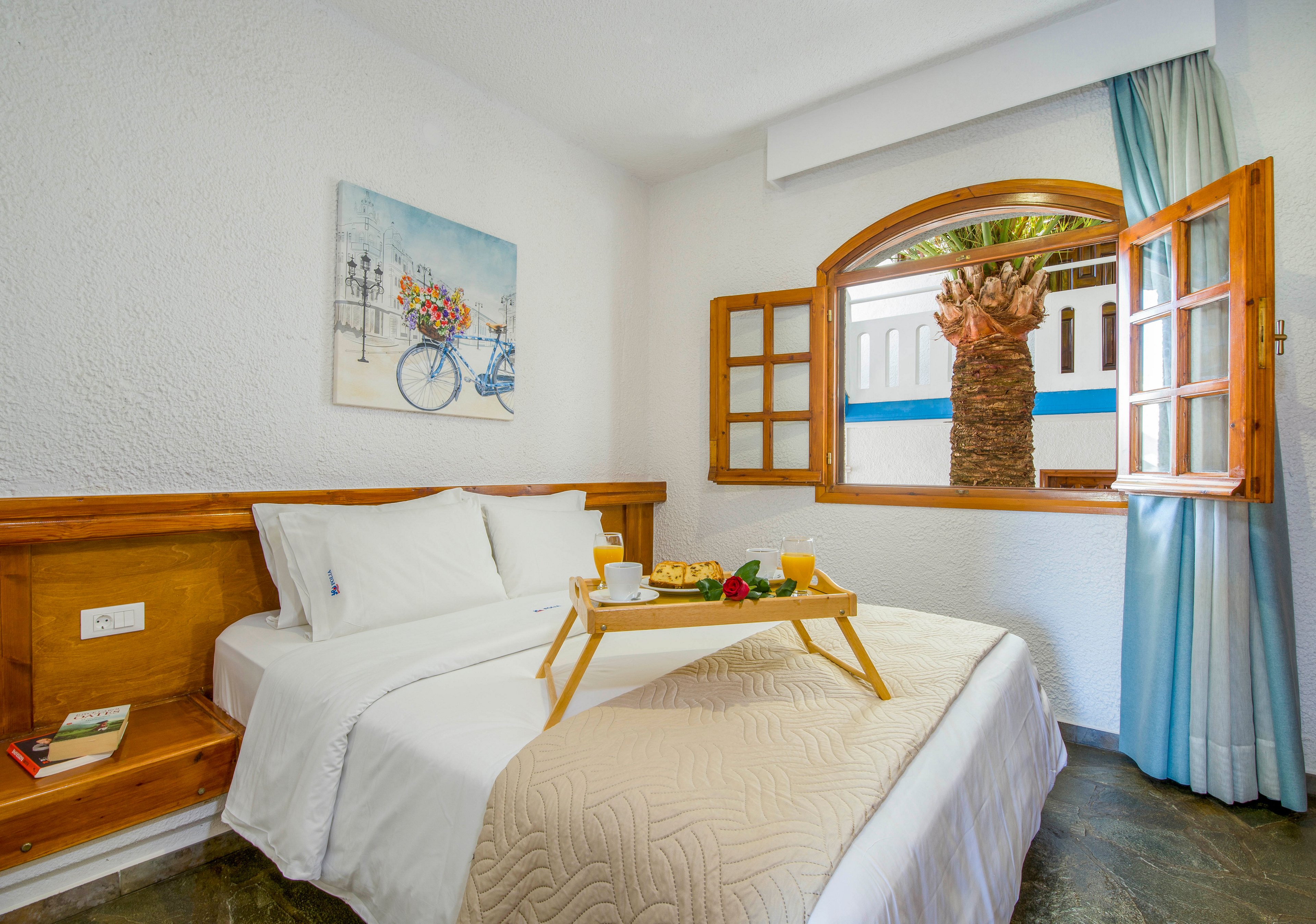 Bright, cozy bedroom with a neatly made double bed and wooden accents. A breakfast tray with orange juice, coffee, and pastries is placed on the bed, adding a welcoming touch. An open window with blue curtains reveals a palm tree and white exterior walls outside. A colorful bicycle painting and a book on the bedside shelf complete the relaxed vacation atmosphere.
