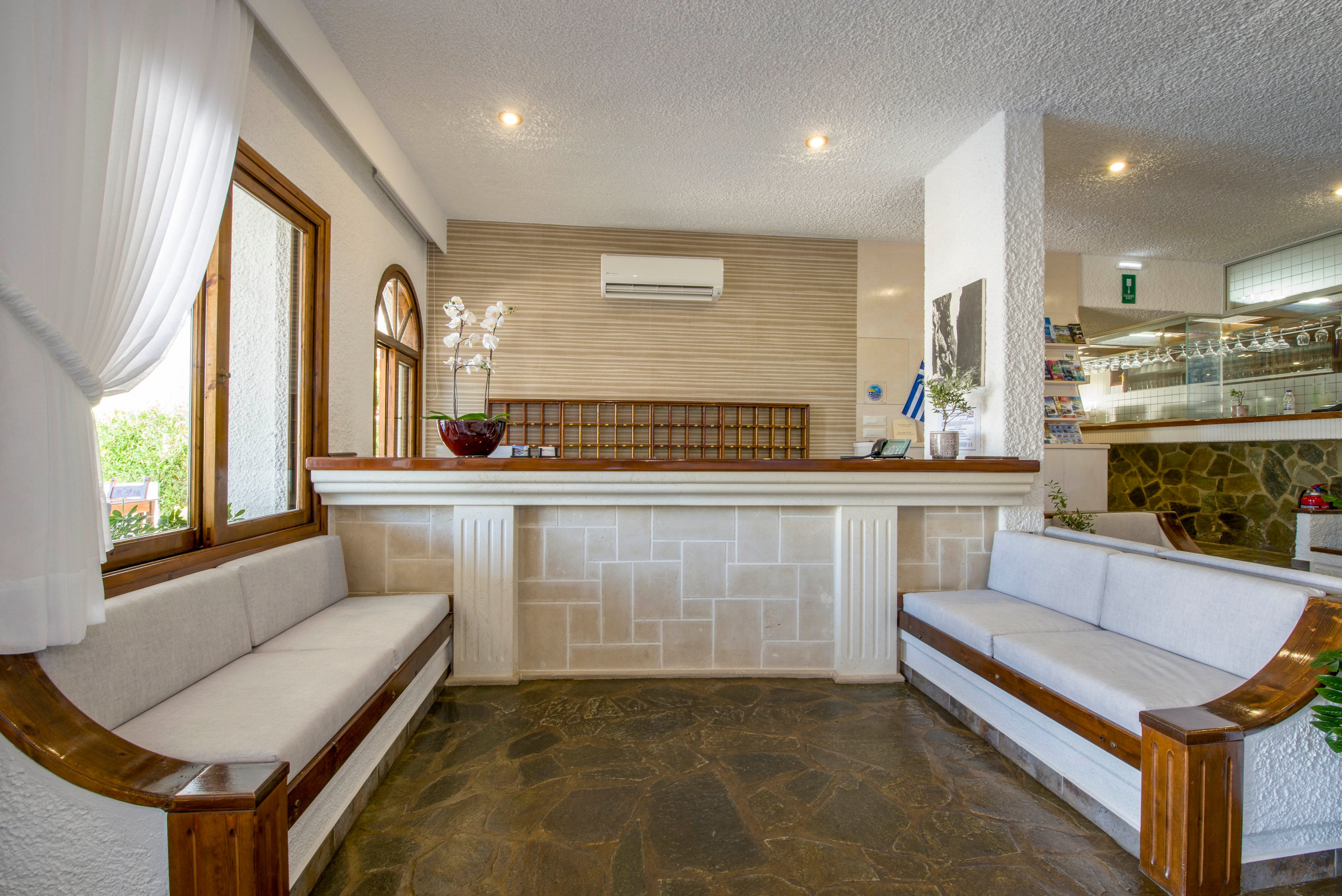 The image shows a clean, bright hotel reception desk with a stone-textured front panel. Two long cushioned benches with wooden frames face the counter, creating a welcoming waiting area. An air conditioning unit is mounted above the desk, and a wall of key slots is visible behind it. Large windows with sheer white curtains bring in natural light, while recessed ceiling lights illuminate the interior.