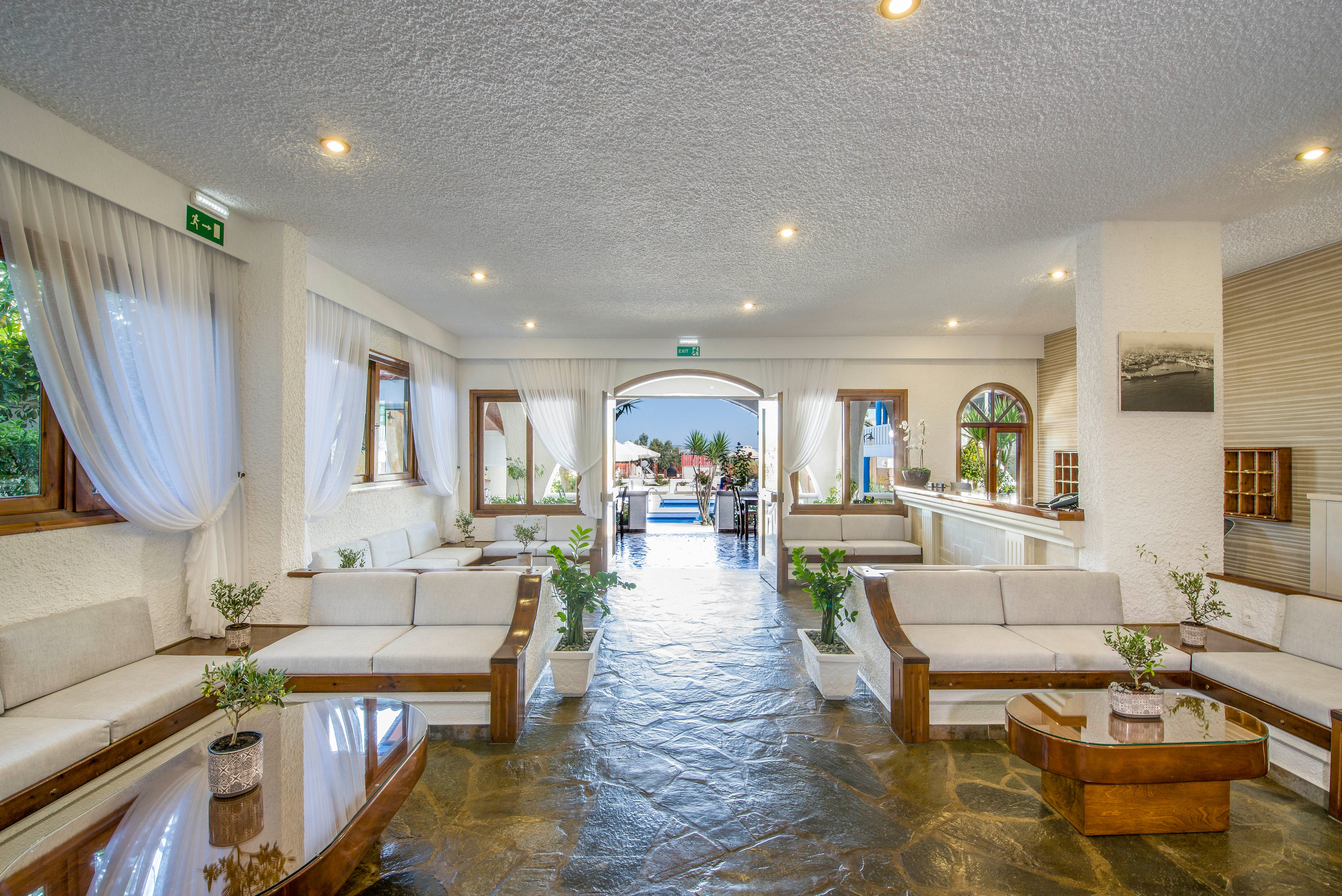 Spacious hotel lobby with white walls and cushioned seating opens toward an outdoor pool area. Large windows with sheer curtains let in natural light and greenery from outside. Wooden tables and planters add warm accents to the stone floor. The scene feels airy, inviting, and relaxed, ideal for vacation or resort imagery.