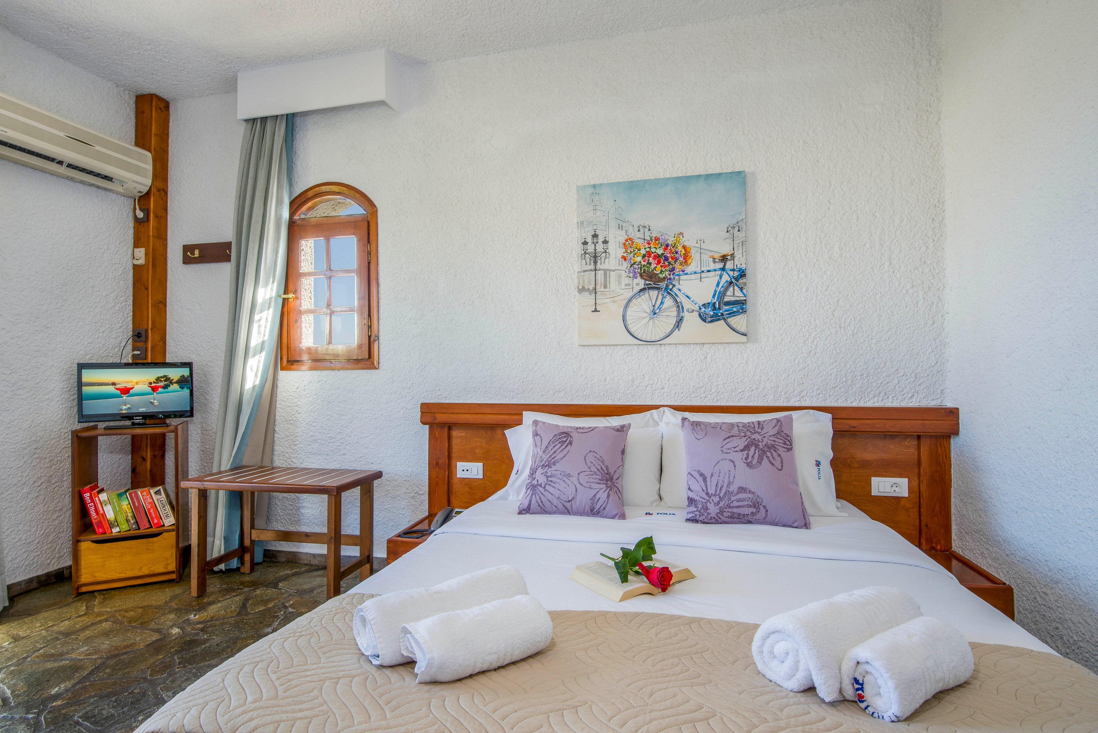 Bright hotel room features a neatly made double bed with white linens and lavender accent pillows. Rolled towels and a small flower arrangement are placed on the bed, adding a welcoming touch. A wooden side table, small bookshelf, and television sit against the wall beneath an air conditioning unit. Textured white walls, a small arched window, and a bicycle artwork create a relaxed Mediterranean atmosphere.