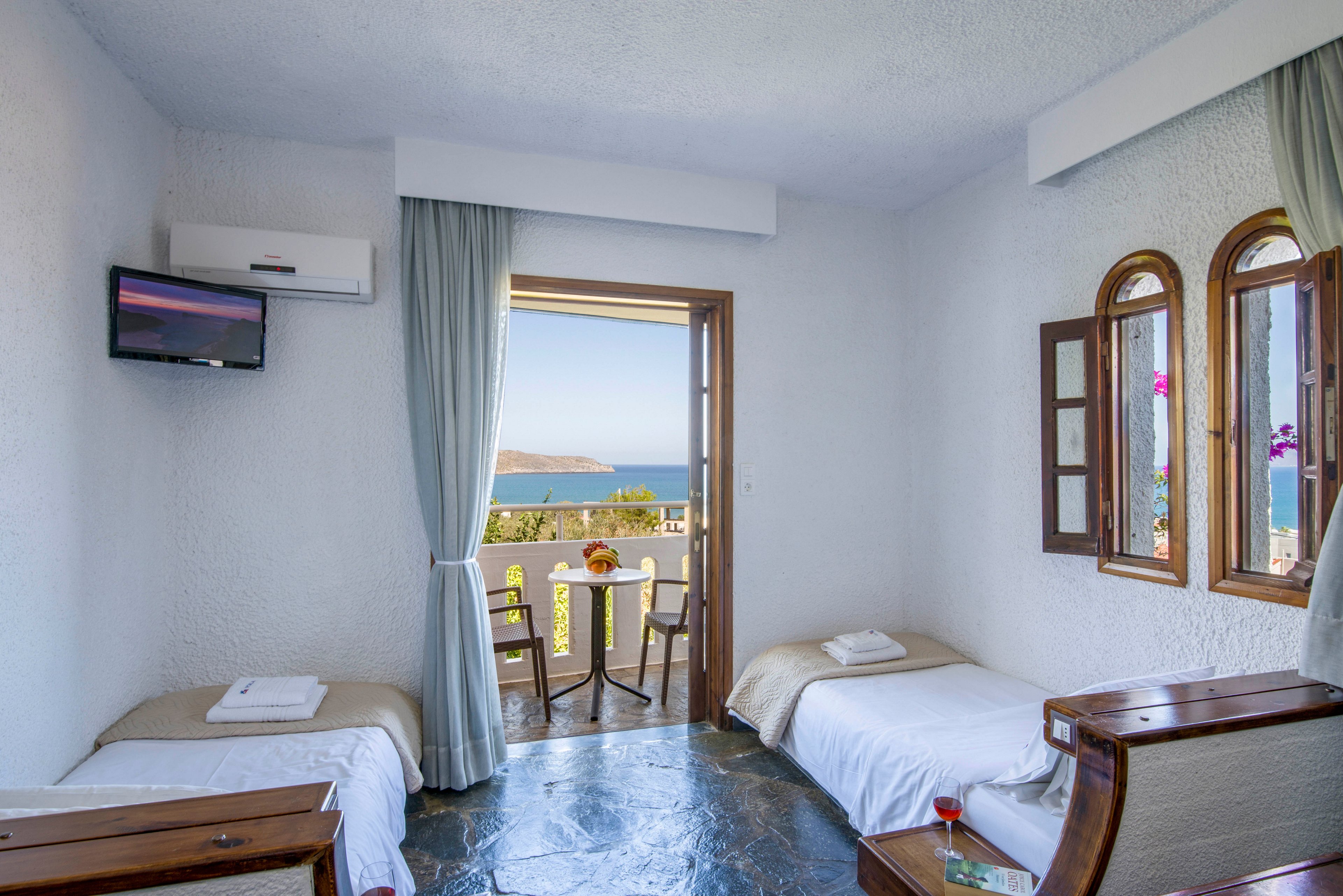 A light-filled hotel room features two single beds with neatly folded white towels. The space opens onto a small balcony with a table and chairs overlooking a calm blue sea and distant coastline. Soft blue curtains frame the doorway, while wooden furniture and arched windows add a warm, Mediterranean touch. A wall-mounted television and air conditioning unit complete the cozy vacation setting.