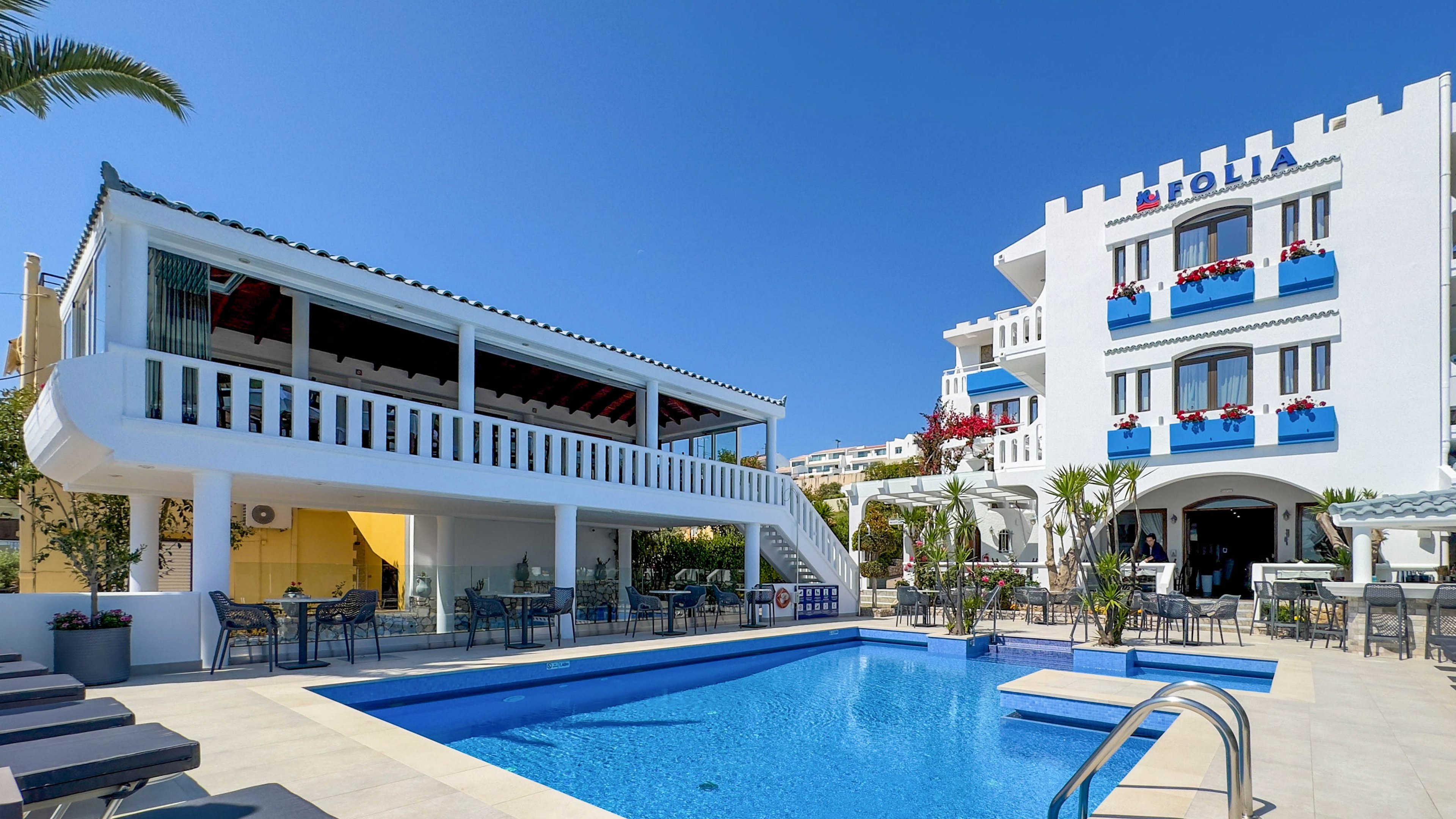A bright white Mediterranean-style hotel surrounds a clear blue outdoor swimming pool under a cloudless sky. The building features blue window shutters, red flowers on balconies, and an elevated terrace with shaded seating. Sun loungers line the pool deck, and palm trees and potted plants add a tropical touch. The scene suggests a relaxing vacation atmosphere in a warm coastal destination.