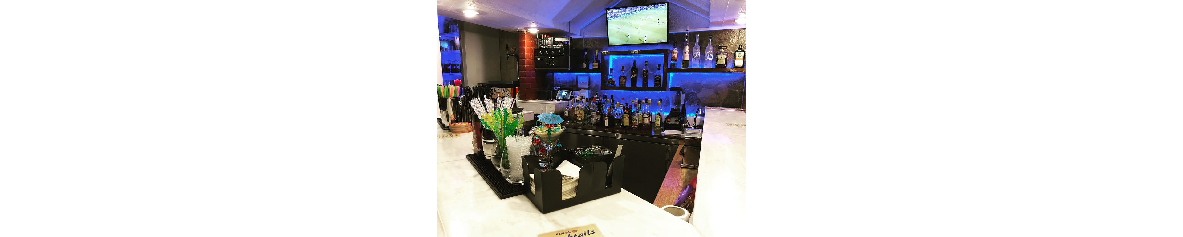 A contemporary bar counter is stocked with assorted liquor bottles and cocktail-making accessories. Colorful stirrers and straws stand beside a glass filled with ice on the white countertop. Blue accent lighting highlights the shelves of spirits in the background. A television mounted above the bar displays a soccer match, adding to the lively atmosphere.