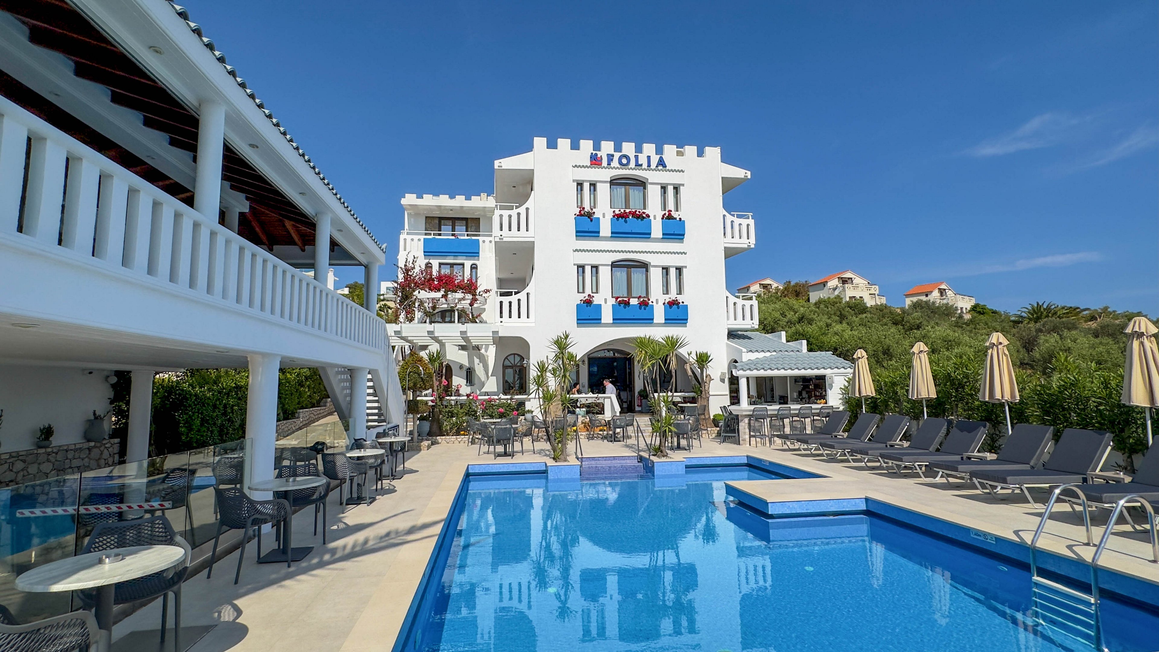 A white Mediterranean-style hotel with blue accents overlooks a clear outdoor swimming pool. Sun loungers and beige umbrellas line the poolside terrace, creating a relaxed vacation atmosphere. The building features balconies with red flowers and surrounding greenery under a bright blue sky. Tables and chairs are arranged along the covered walkway, suggesting an inviting space for guests to sit and enjoy the view.
