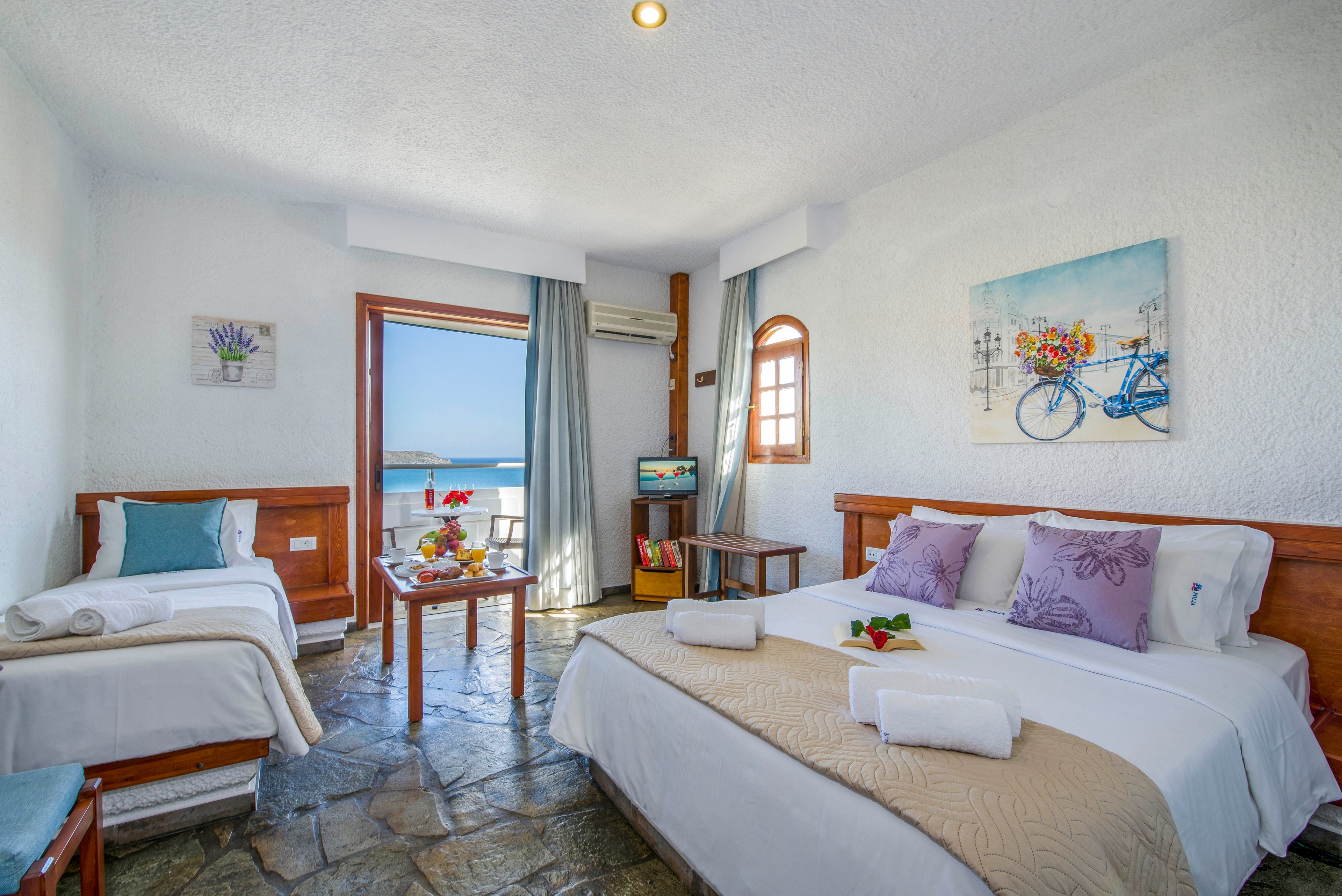 A spacious, sunlit hotel room features a large double bed and a single bed with neatly folded white towels. Stone flooring and wooden furniture create a rustic, coastal feel, complemented by light-colored walls and soft pastel pillows. A small table near the open balcony door holds fruit and drinks, framing a clear view of the blue sea outside. Wall art with flowers and a bicycle adds a cheerful decorative touch to the serene vacation setting.