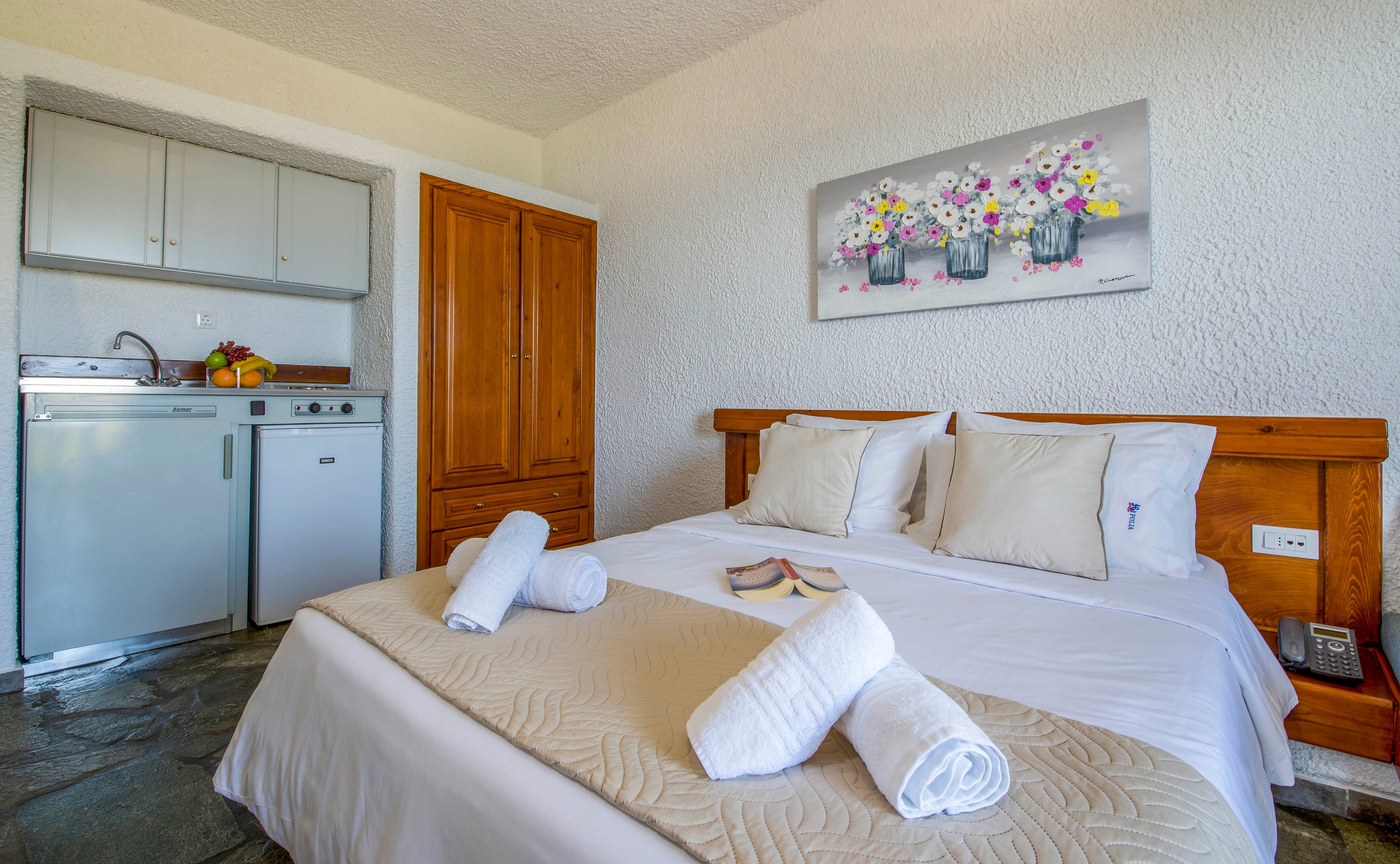 A neatly made double bed with white linens and rolled towels is the focal point of a compact studio room. Beside the bed, a small kitchenette with a sink, stovetop, and fruit bowl provides basic cooking facilities. A wooden wardrobe stands between the kitchenette and the bed, adding warm tones to the otherwise light interior. Above the headboard, a colorful floral painting brightens the space, creating a welcoming and relaxed atmosphere.