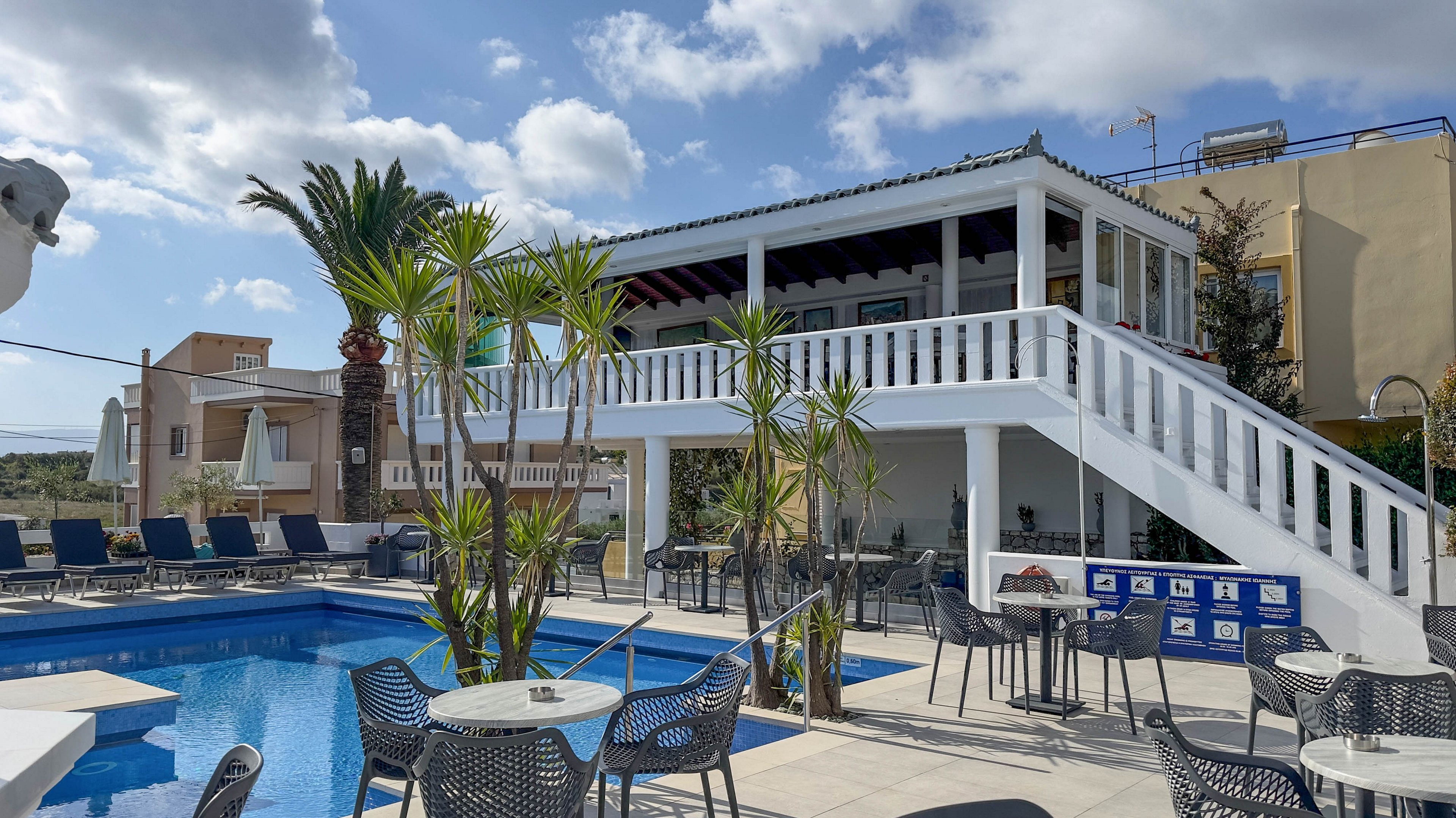 An outdoor hotel pool area features a bright blue swimming pool surrounded by lounge chairs and café tables. A white two-story terrace with stairs overlooks the pool, creating a relaxed vacation setting under a partly cloudy sky. Tall palm trees and potted plants add greenery to the scene, while nearby buildings frame the courtyard. The atmosphere suggests a warm, sunny day ideal for leisure and socializing by the water.