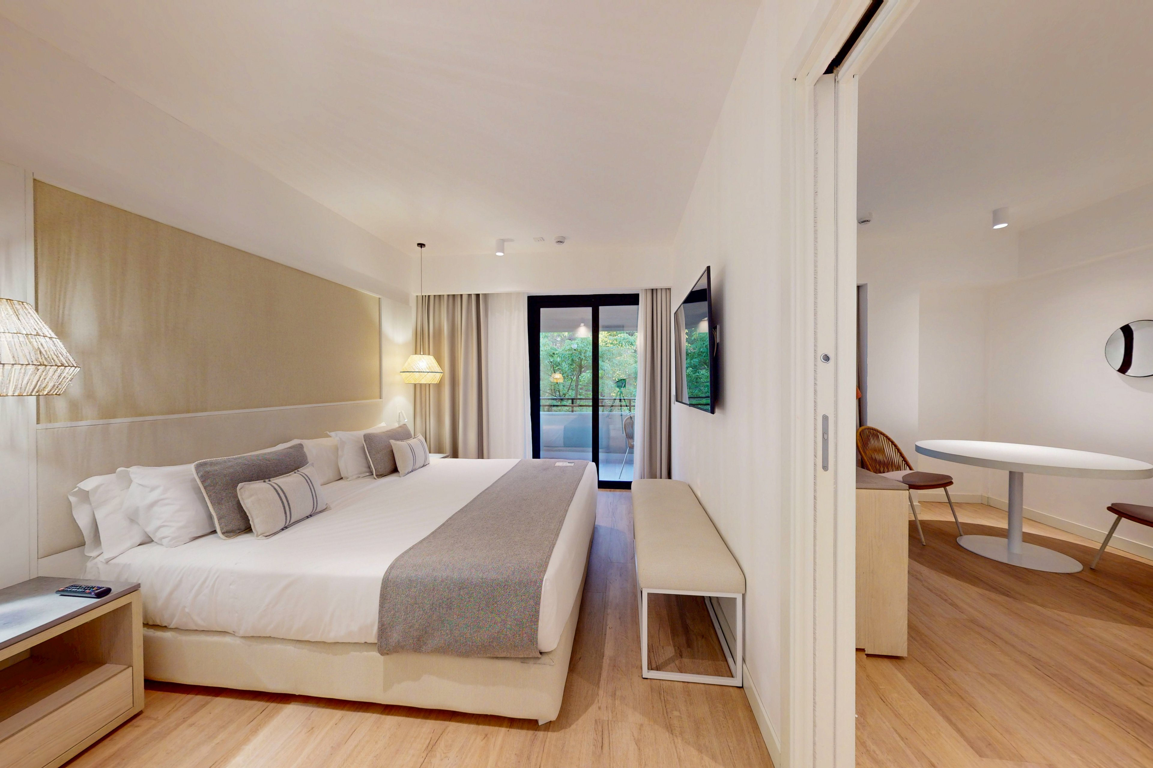 A spacious and contemporary hotel room featuring a large bed with neutral-toned bedding and decorative pillows. The setting includes a wooden floor, minimalist furniture, and a sliding door leading to a balcony with greenery visible outside. Warm lighting and clean design create a cozy and inviting atmosphere.