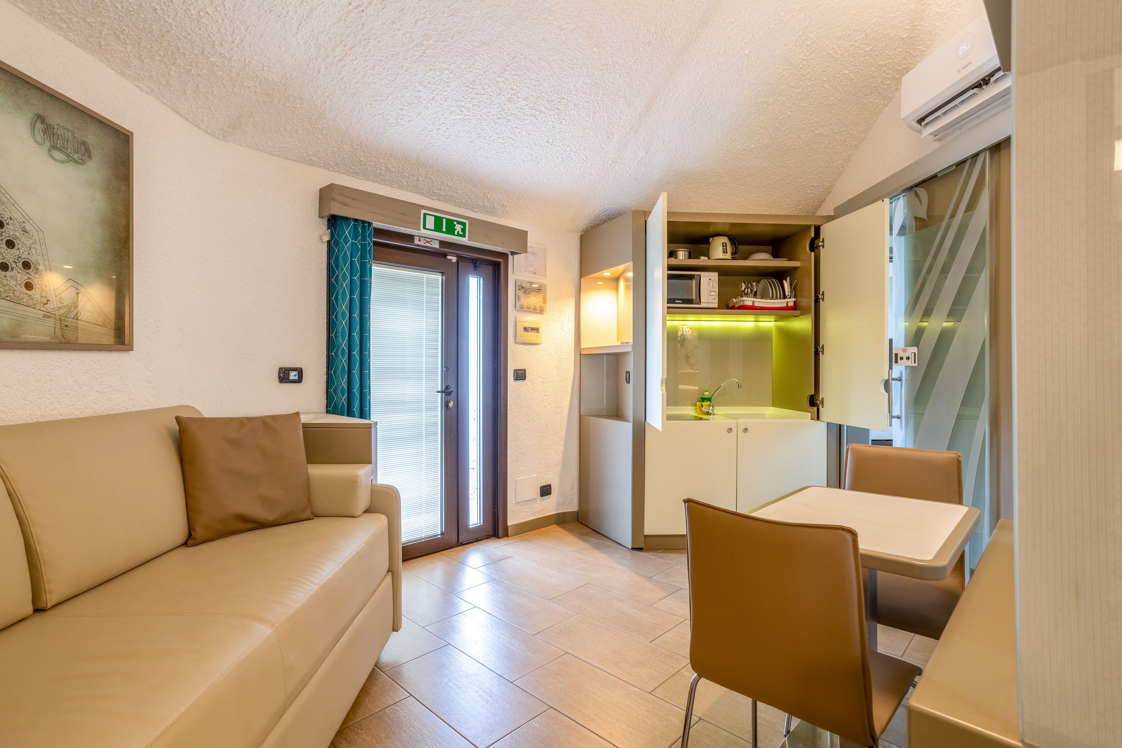 Bright studio interior featuring a beige sofa and small dining table with two chairs. A compact kitchenette with open cabinets, microwave, and under-cabinet lighting sits against one wall. Natural light enters through a glass door with blue curtains and an overhead emergency exit sign. Neutral tones and clean lines create a modern, welcoming atmosphere suitable for short stays or rentals.