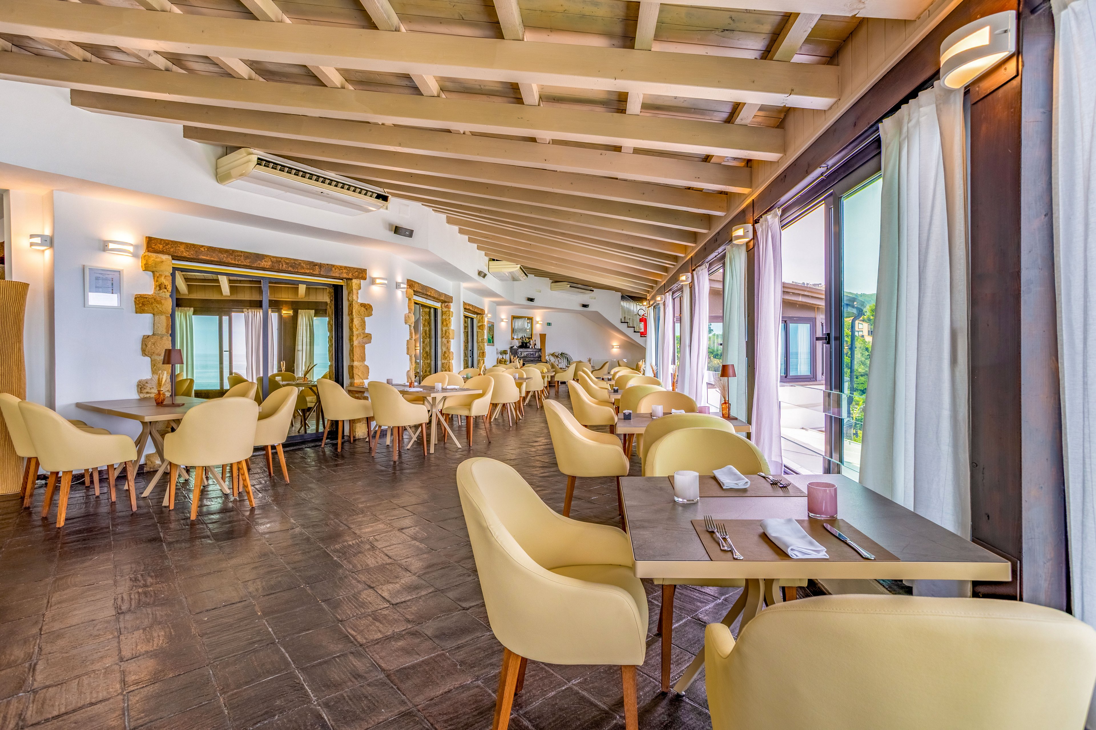 Spacious contemporary restaurant dining area with light yellow chairs and wooden tables is shown in daylight. The room features large windows with sheer curtains, offering views of an outdoor terrace and greenery. Exposed wooden ceiling beams and tiled flooring create a warm, rustic-modern atmosphere. Tables are neatly set with napkins, cutlery, and small decorative items, suggesting readiness for guests.