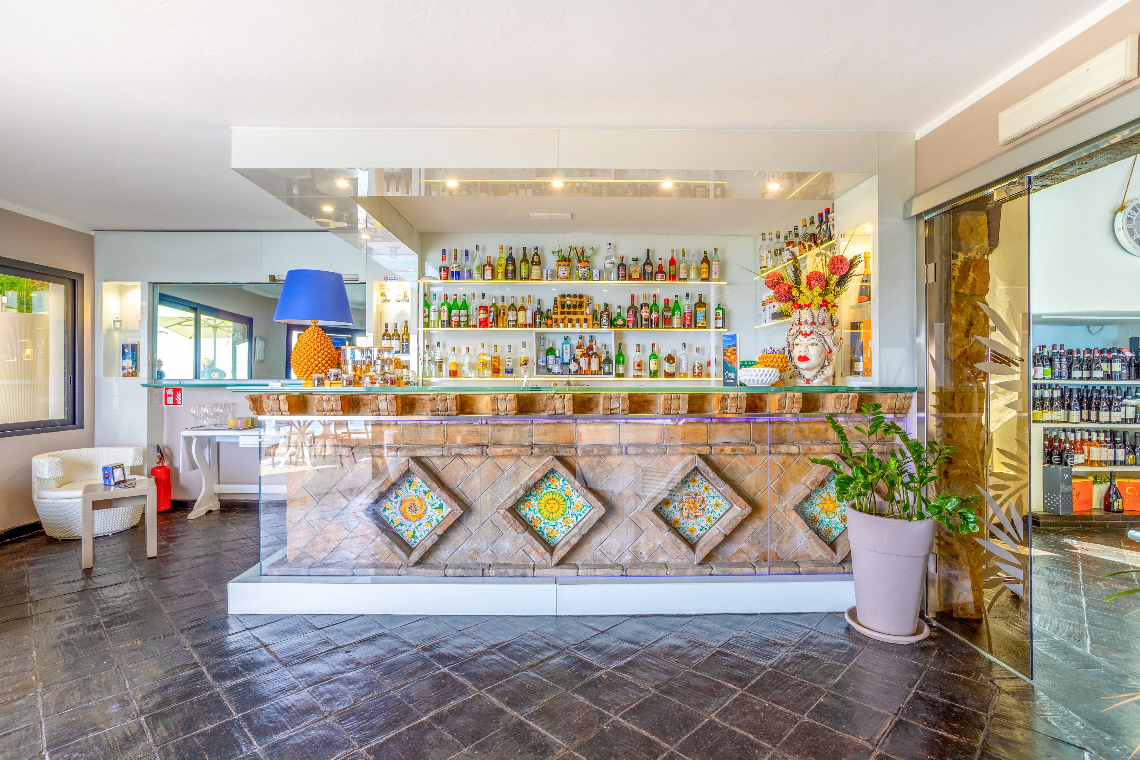 A modern hotel bar features a long counter decorated with colorful patterned tiles and glass shelving filled with assorted bottles. The space is bright and airy, with large windows, white walls, and contemporary seating in the foreground. A blue table lamp, decorative vases, and a floral arrangement add vibrant accents to the interior. The polished floor and mirrored surfaces create reflections that enhance the spacious, welcoming atmosphere.