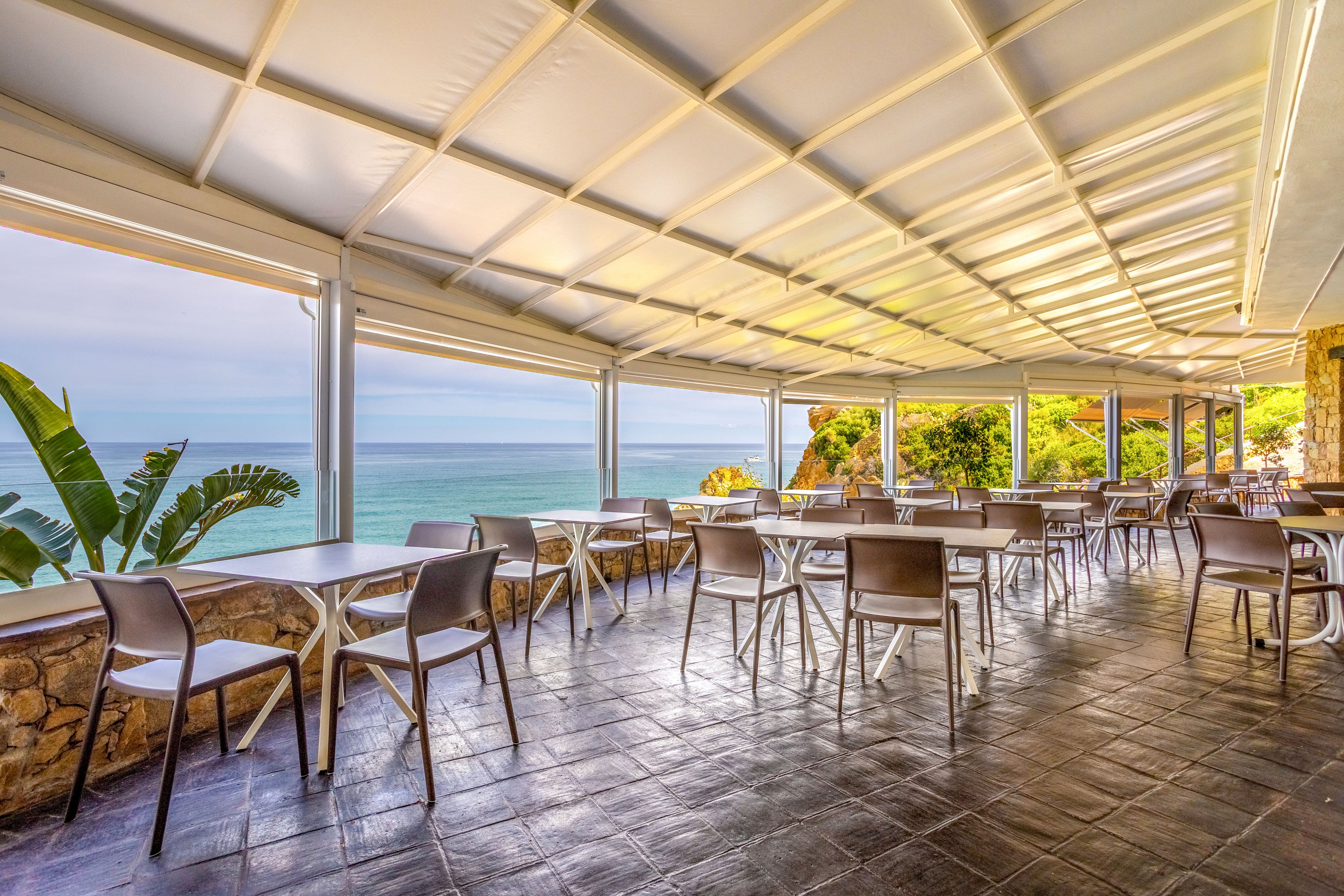 An open-air restaurant terrace overlooks a calm blue ocean under a covered roof structure. Empty tables and chairs are neatly arranged on a tiled floor, suggesting a peaceful, off-peak moment. Large openings frame the sea and sky, while lush green foliage and rocky walls add a natural coastal atmosphere. The bright daylight and clean setting create a relaxed, inviting mood for outdoor dining by the water.