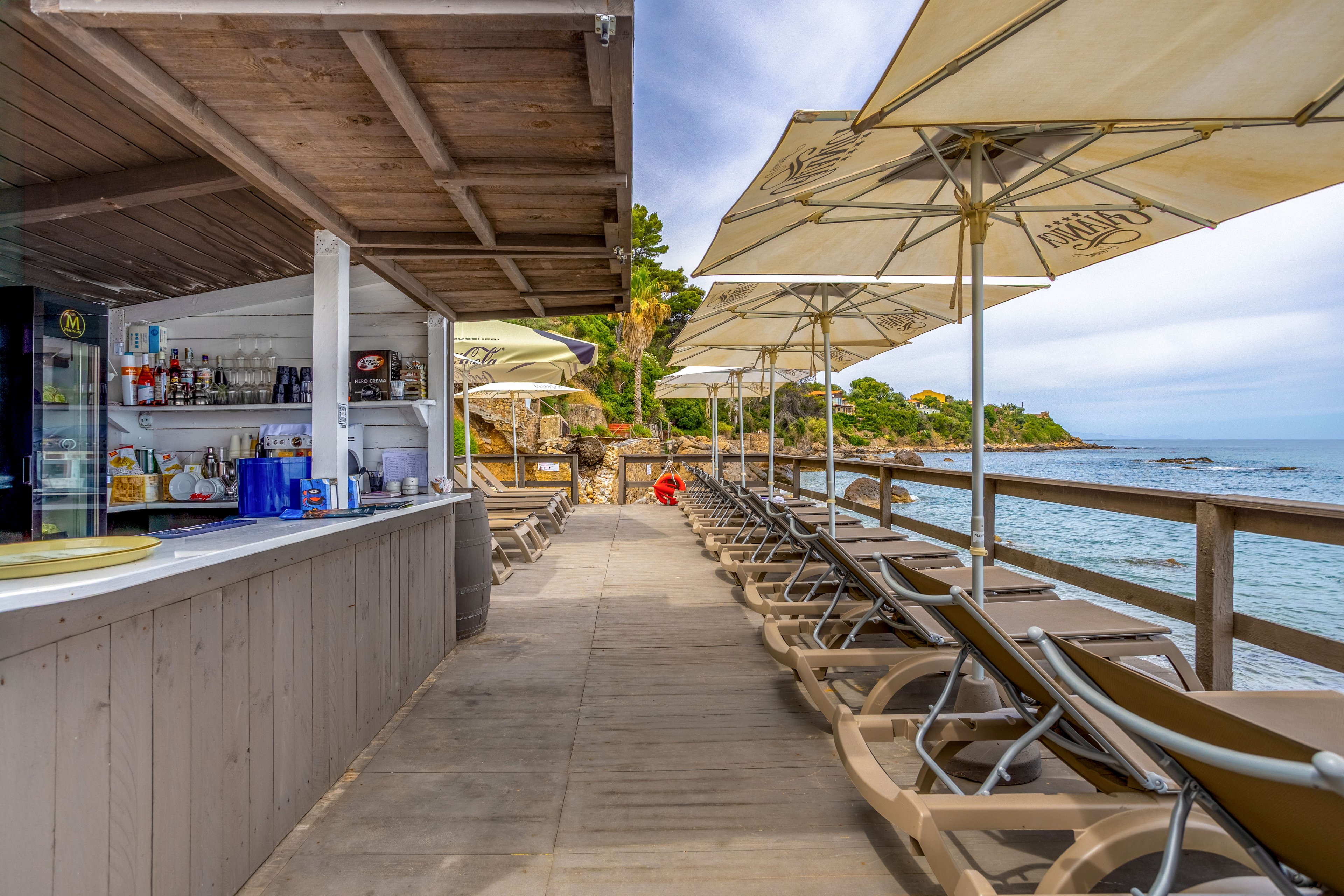 An open-air beach bar stands beside a row of empty sun loungers under large umbrellas on a wooden deck. The scene overlooks a calm blue sea with rocky shoreline and green trees in the background. Bottles, glasses, and a refrigerator are visible behind the bar counter, suggesting refreshments for visitors. The overall atmosphere is relaxed and inviting, ideal for vacation or summer leisure by the water.