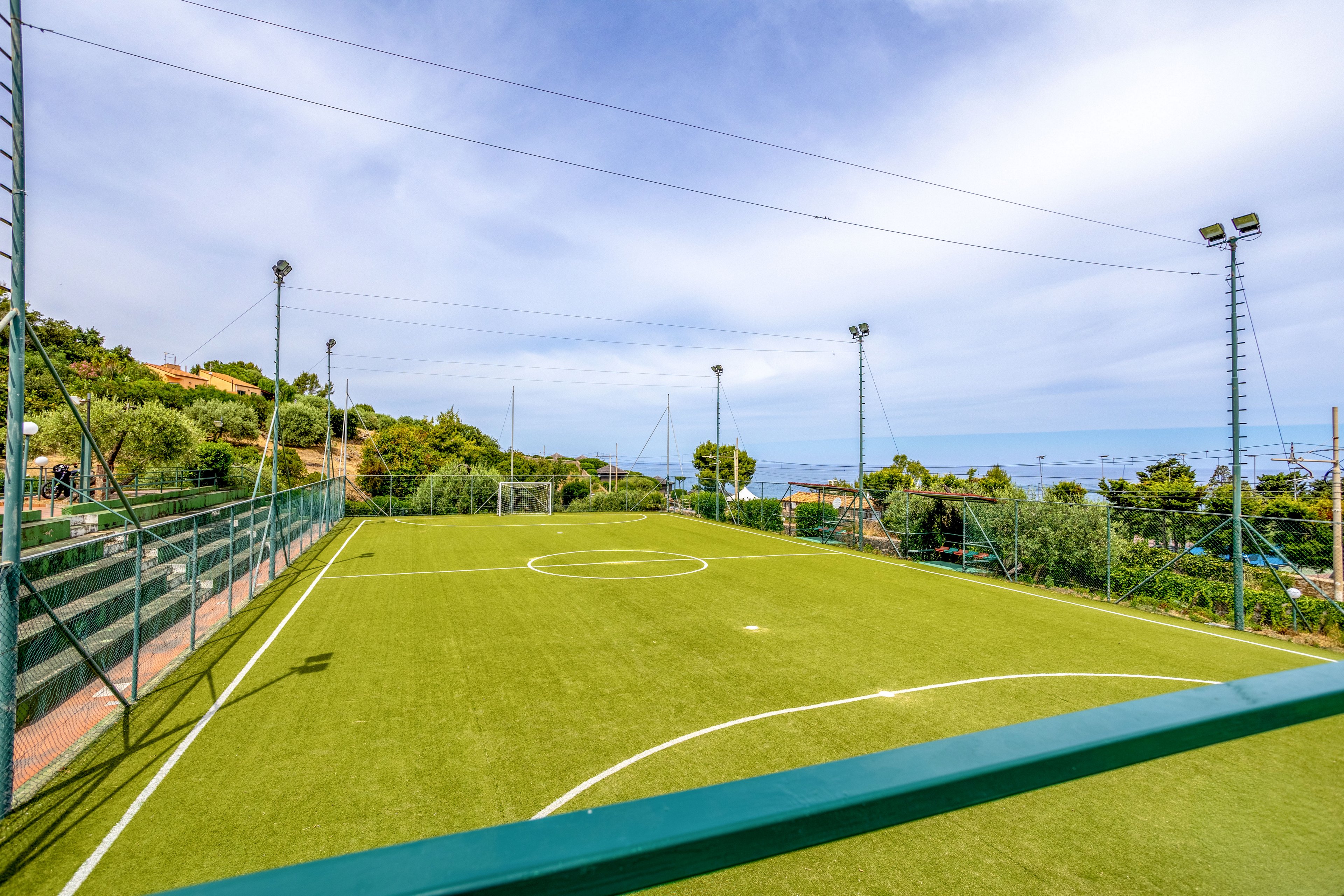An empty artificial turf soccer field is shown under a bright blue sky. The pitch is surrounded by tall fencing and lighting poles, with small goals visible at each end. Lush greenery and hillside buildings frame the scene, and the sea can be seen in the distance. The overall atmosphere is calm and inviting, suggesting a recreational sports area near the coast.