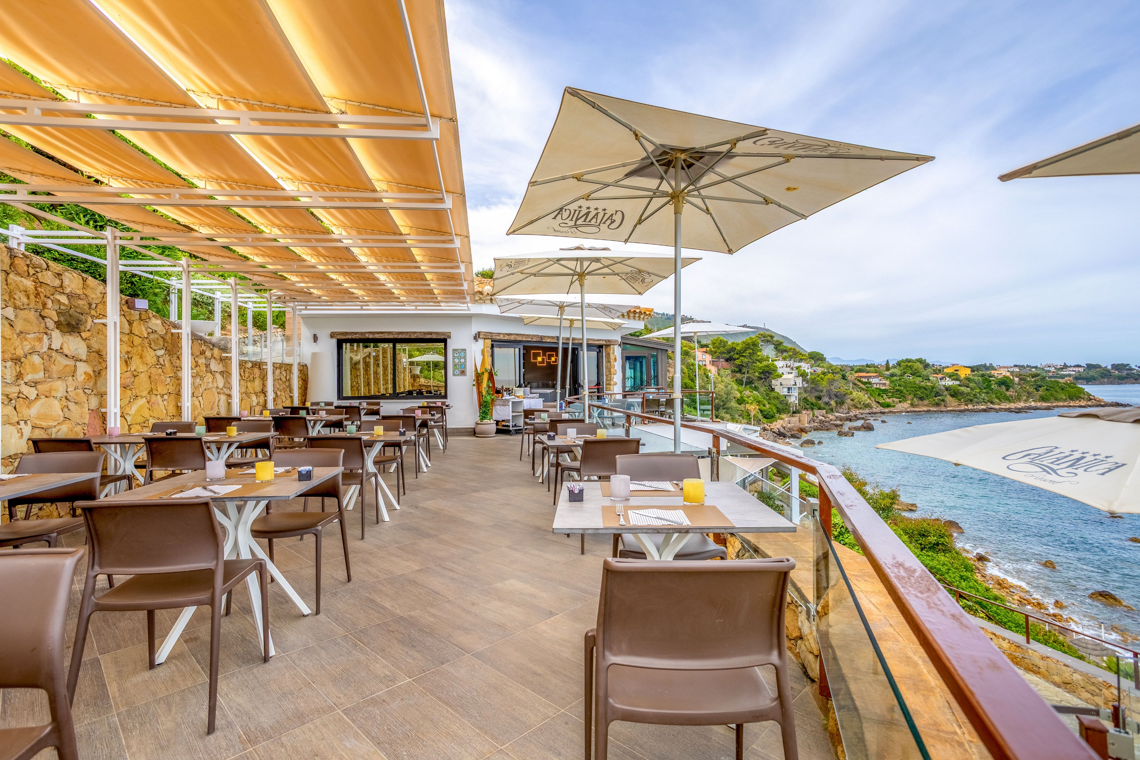 An outdoor restaurant terrace overlooks a rocky coastline and calm sea. Brown chairs and metal tables are neatly arranged under large cream-colored sun umbrellas and retractable awnings. A stone wall and glass railing frame the dining area, while distant houses and greenery line the shore. The bright daylight and clear sky create a relaxed, inviting atmosphere for al fresco dining.