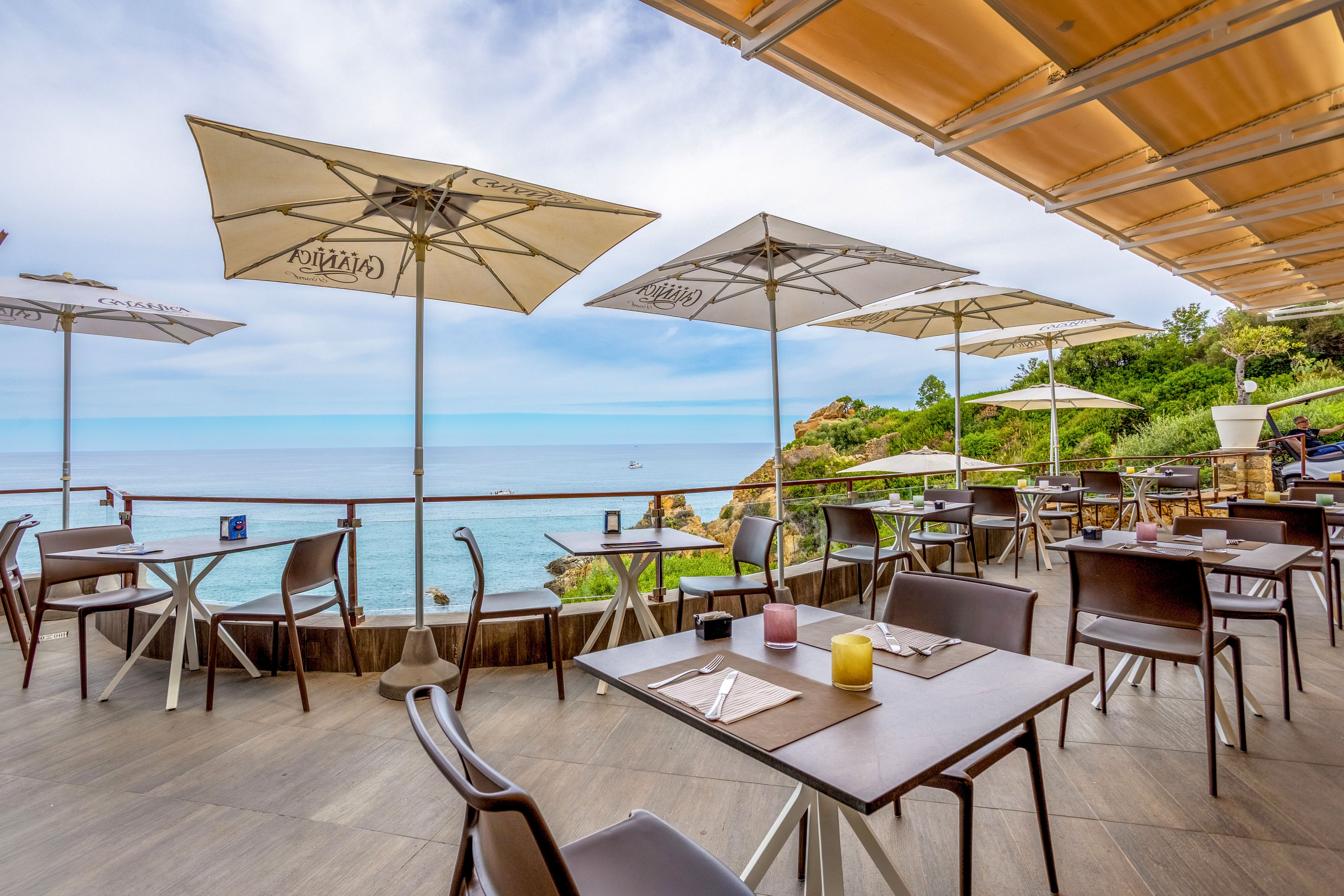 An open-air café terrace overlooks a calm blue sea under a partly cloudy sky. Empty tables and chairs are neatly arranged, each set with placemats, cutlery, and small candles or containers. Large white umbrellas provide shade, and a railing separates the seating area from the water below. Greenery and rocky cliffs frame the scene, creating a relaxed coastal atmosphere.