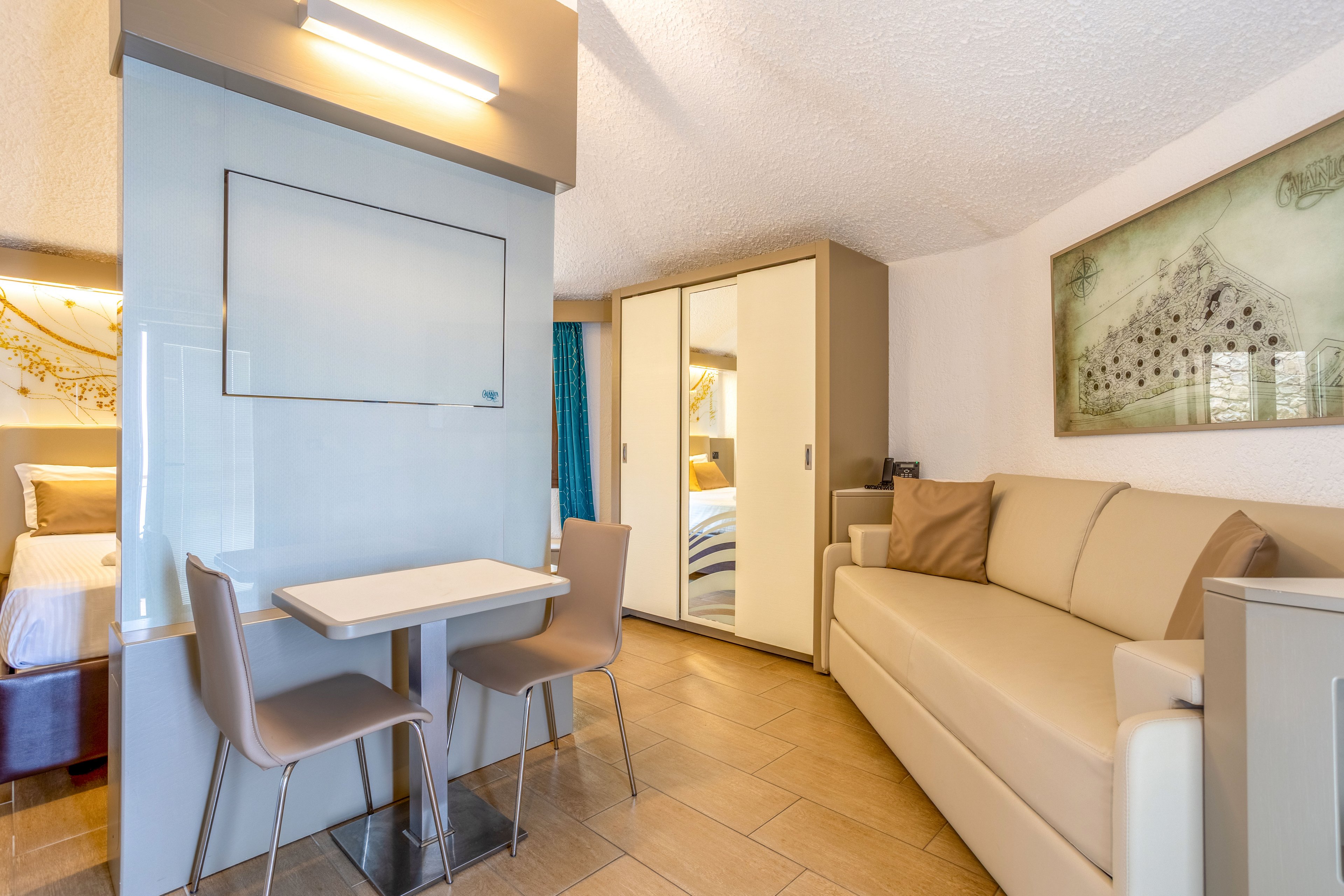 Contemporary studio apartment featuring a beige sofa and compact dining area with two chairs and a small table. A white wardrobe with sliding doors separates the living space from the sleeping area, where a bed is partially visible. Warm lighting and neutral tones create a cozy, inviting atmosphere. A framed artwork hangs above the sofa, adding decorative interest to the clean, minimalist design.