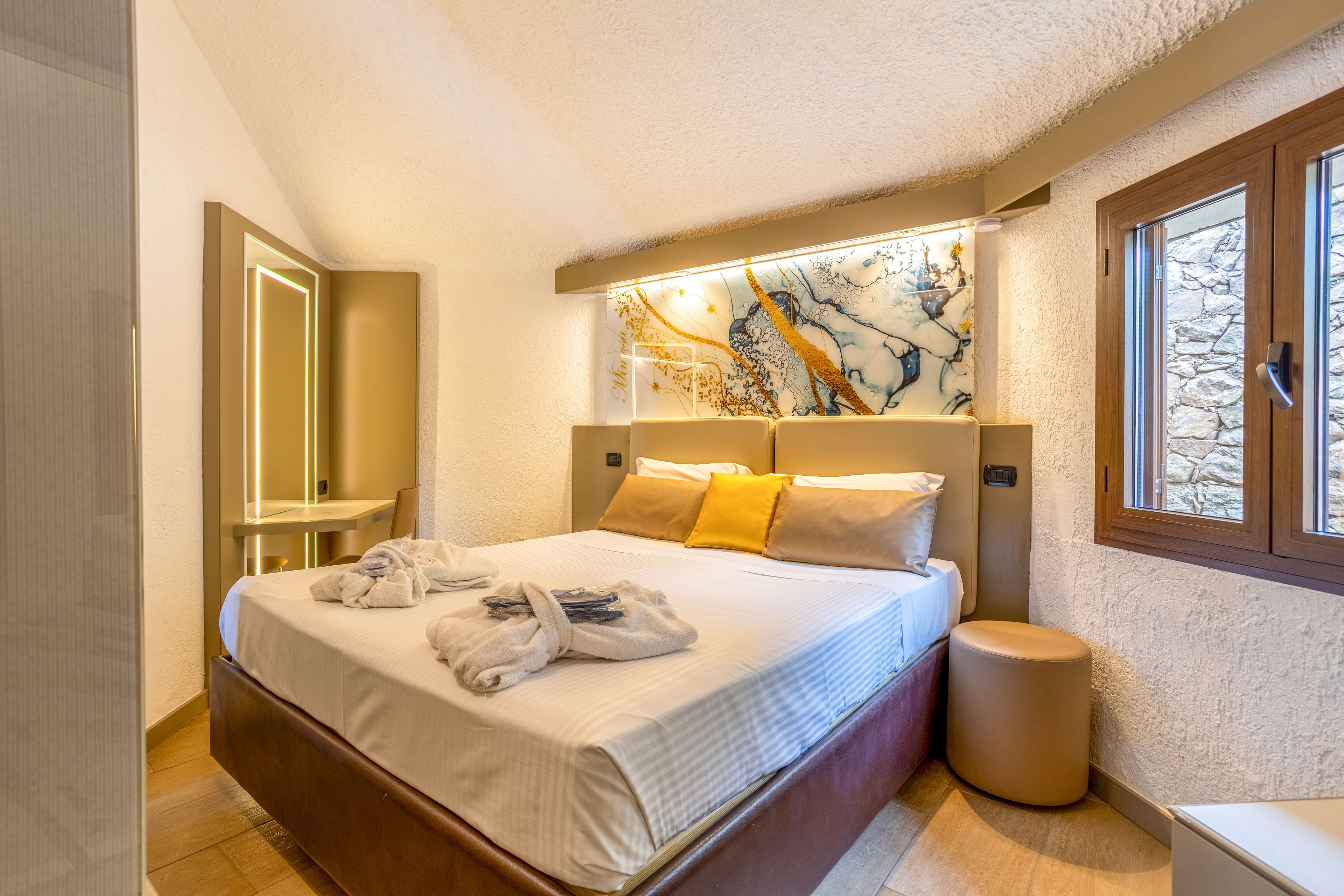 A neatly made double bed with white linens and folded bathrobes is the focal point of this modern hotel bedroom. Warm ambient lighting highlights a decorative abstract wall panel above the headboard, adding artistic flair to the space. A small desk with an illuminated mirror stands to the left, while a wooden-framed window on the right lets in natural light. Soft neutral tones with yellow accents create a welcoming and comfortable atmosphere.