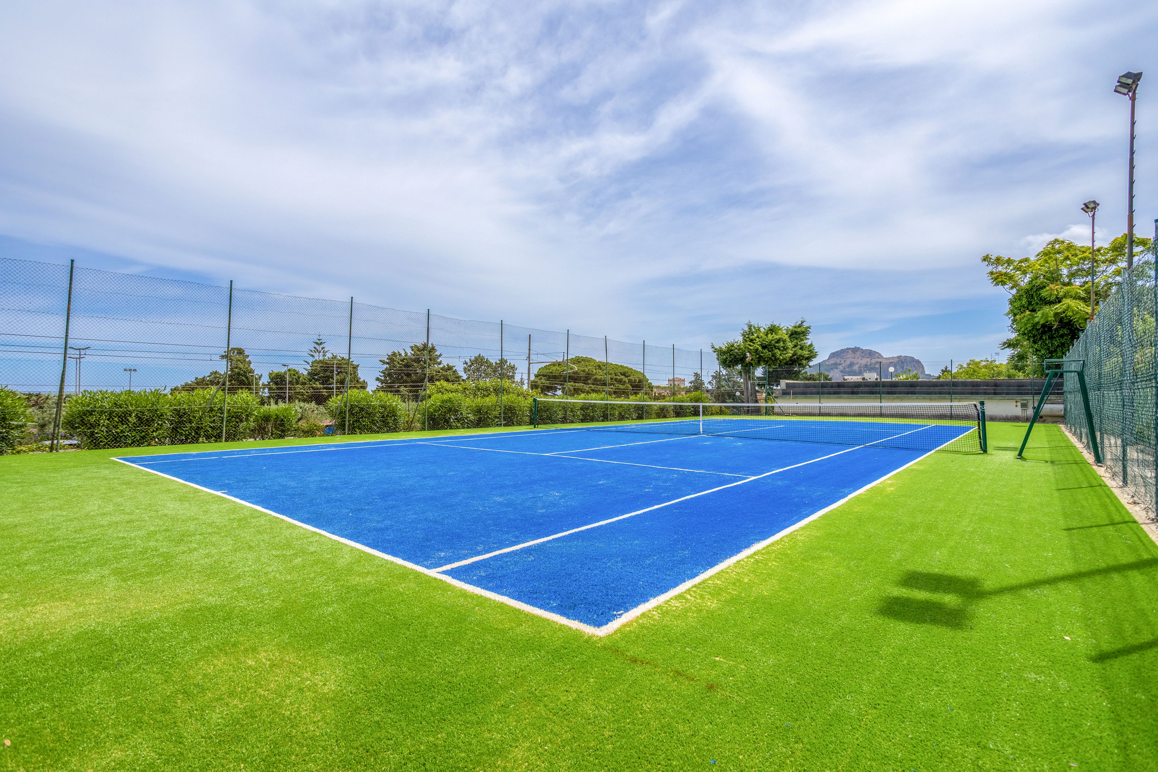 A bright blue tennis court sits empty in an outdoor sports facility. The court is surrounded by vivid green artificial grass and tall perimeter fencing. Trees, shrubs, and distant hills frame the background under a partly cloudy sky. The scene suggests a calm, sunny day ideal for tennis or recreational activities.