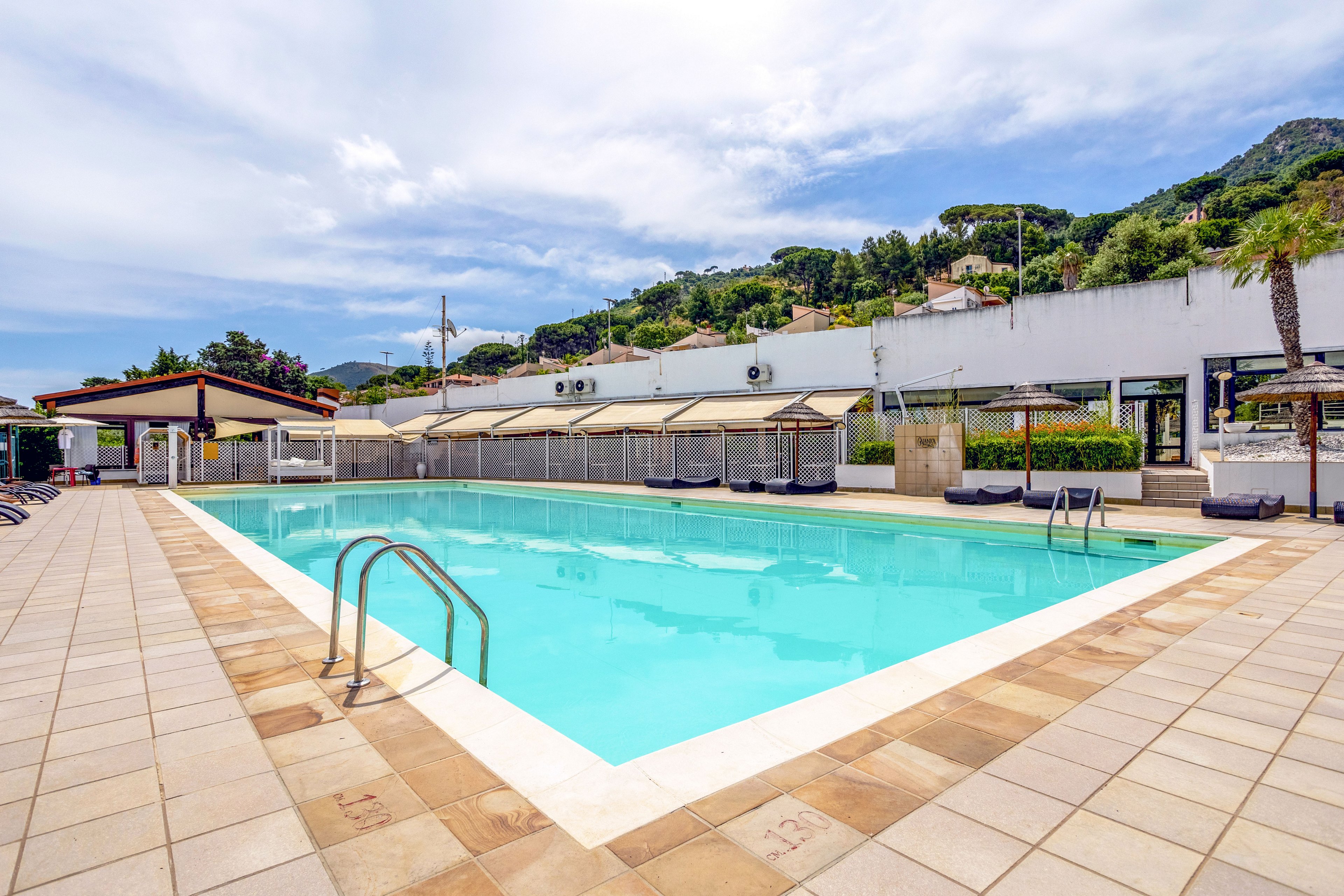 An empty rectangular swimming pool sits in an open-air terrace area on a bright, sunny day. Metal ladders descend into the clear blue water, surrounded by a tiled deck with lounge chairs and shaded cabanas. In the background, white buildings and structures line the edge of the pool area, with green hills rising behind them under a partly cloudy sky. The overall scene suggests a peaceful vacation setting focused on relaxation and leisure.