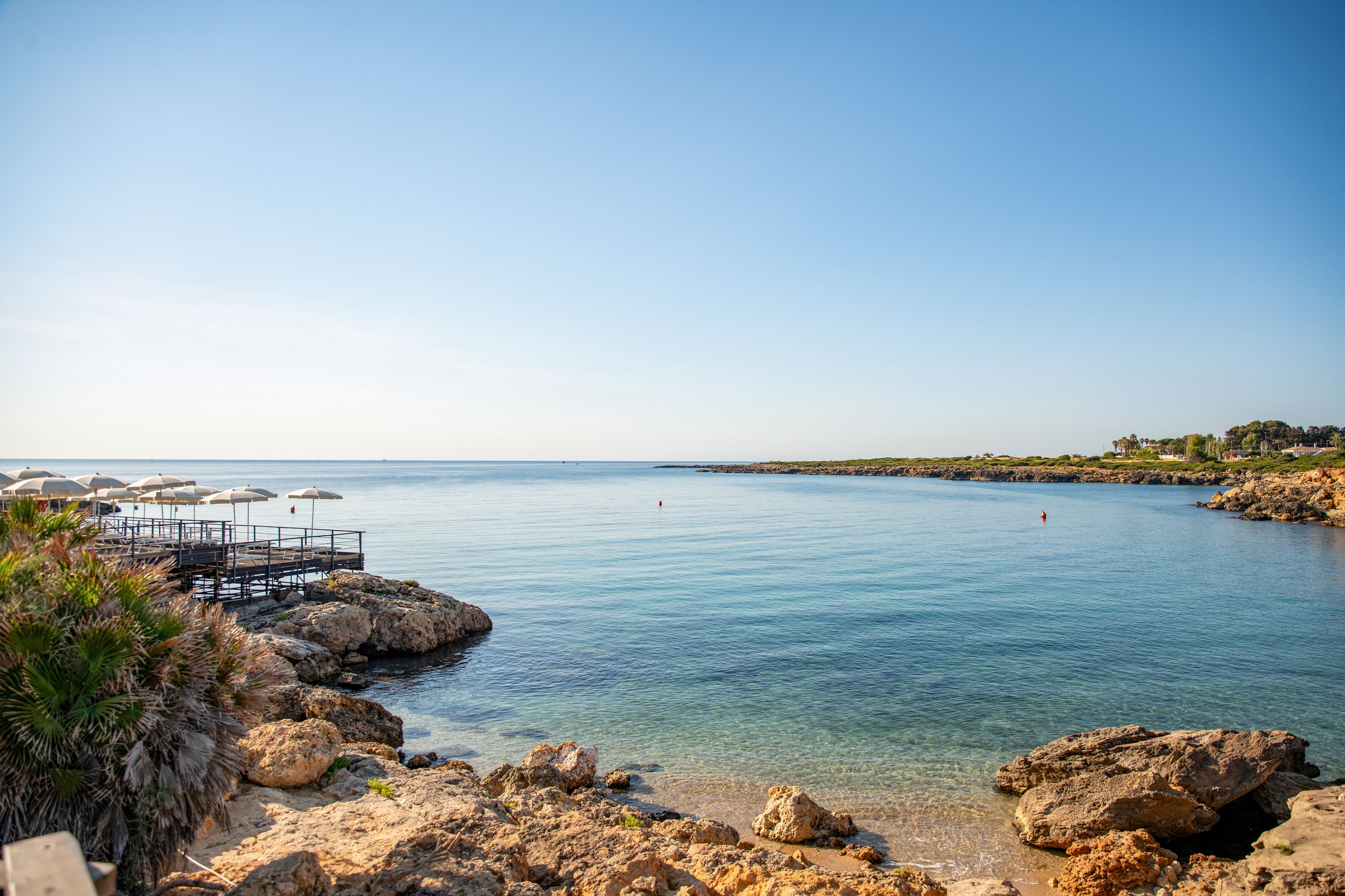 A tranquil coastal bay features clear, calm blue water framed by a rocky shoreline. A small wooden deck with white sun umbrellas extends over the water on the left side, suggesting a seaside resort or beach club. The sky is bright and cloudless, creating a serene and sunny atmosphere. Low vegetation and palm-like plants grow among the rocks, adding a touch of greenery to the coastal scene.