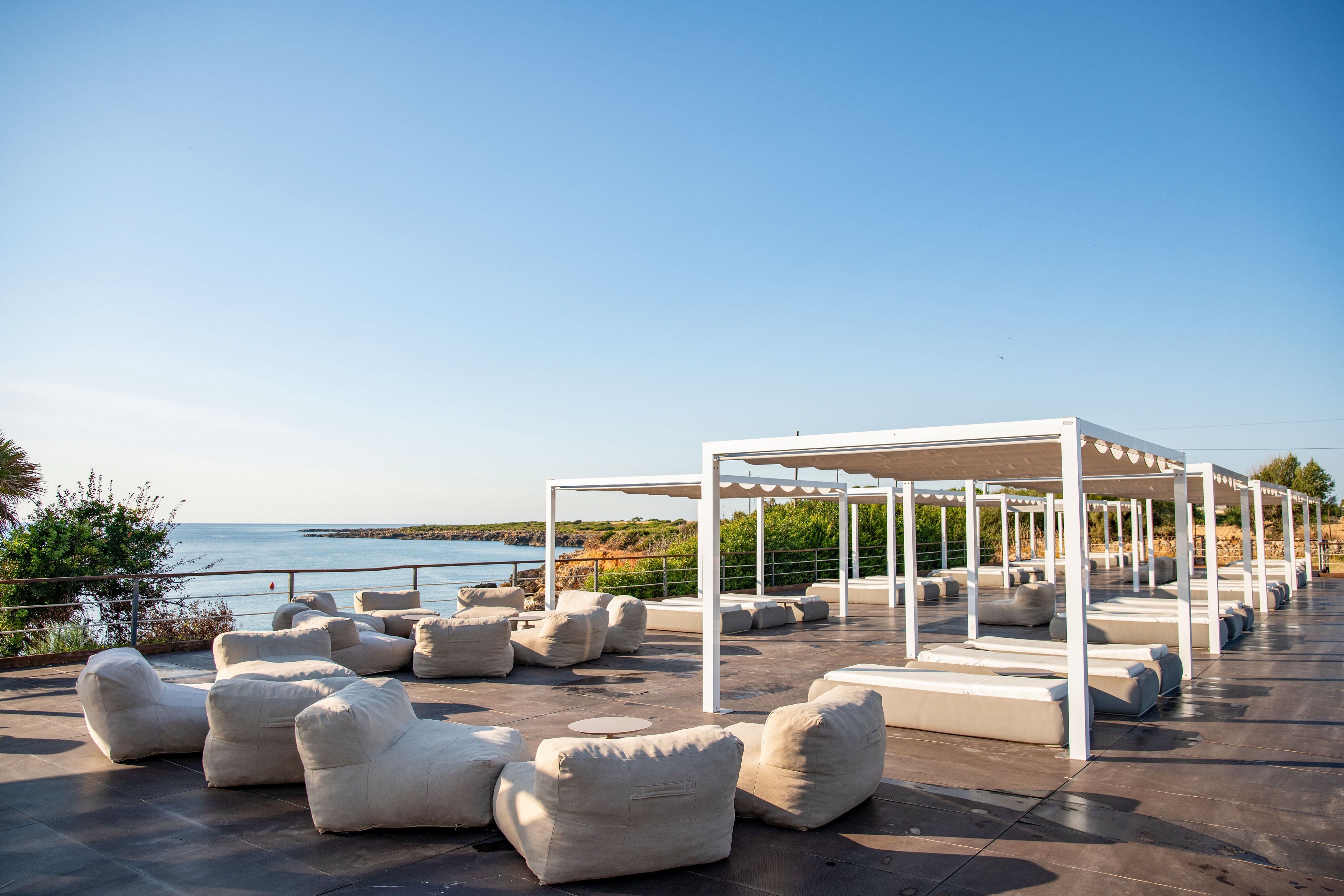 An open-air seaside terrace features white pergola-style canopies and numerous cushioned lounge seats arranged on a wide paved deck. The setting overlooks a calm blue sea and a rocky shoreline with greenery in the background. The sky is clear and bright, suggesting a sunny, relaxing day by the water. The overall scene conveys a modern, minimalist outdoor leisure space designed for comfort and coastal views.