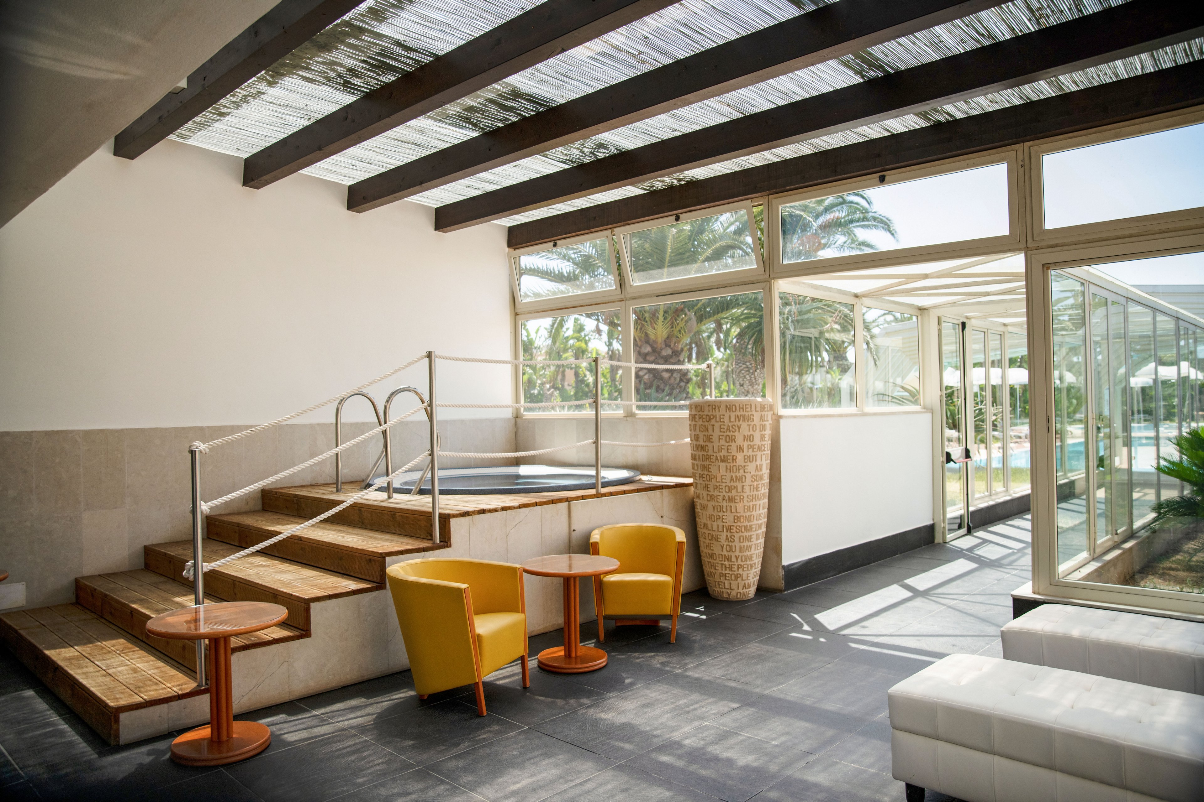 Bright contemporary lounge area with two yellow armchairs and small round tables near a raised indoor pool or hot tub. Large windows and glass doors open to an outdoor space with palm trees and greenery. Wooden beams and neutral walls create a clean, modern aesthetic, complemented by a tall woven floor vase. Natural light fills the room, highlighting the relaxed, resort-like atmosphere.
