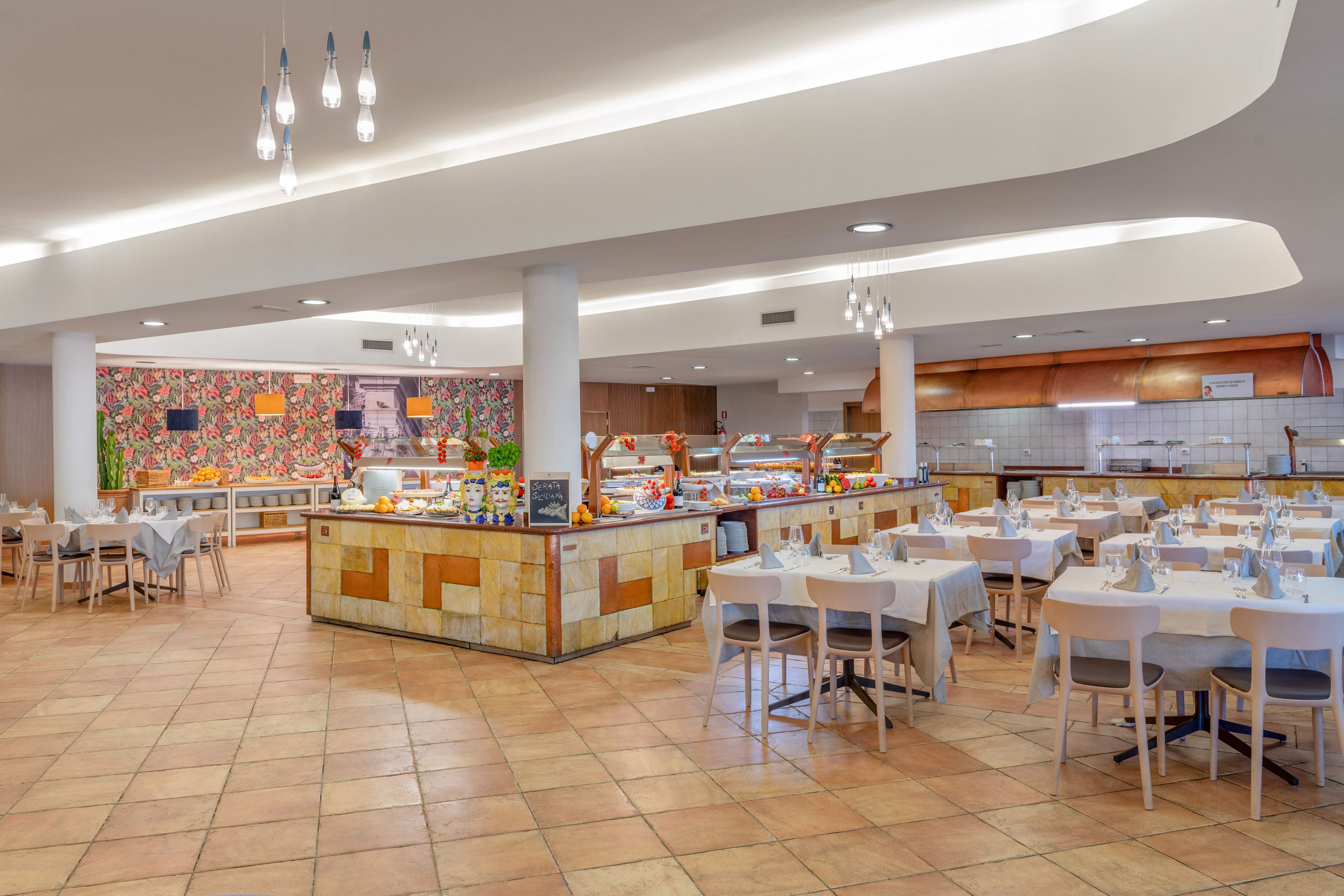 Spacious self-service restaurant interior featuring a central buffet counter and neatly arranged dining tables. White tablecloths and light wooden chairs create a clean, modern look across the tiled floor. The buffet area is brightly lit, with colorful food displays and a decorative wall in the background. Ceiling lights and curved architectural elements add a contemporary touch to the welcoming dining space.