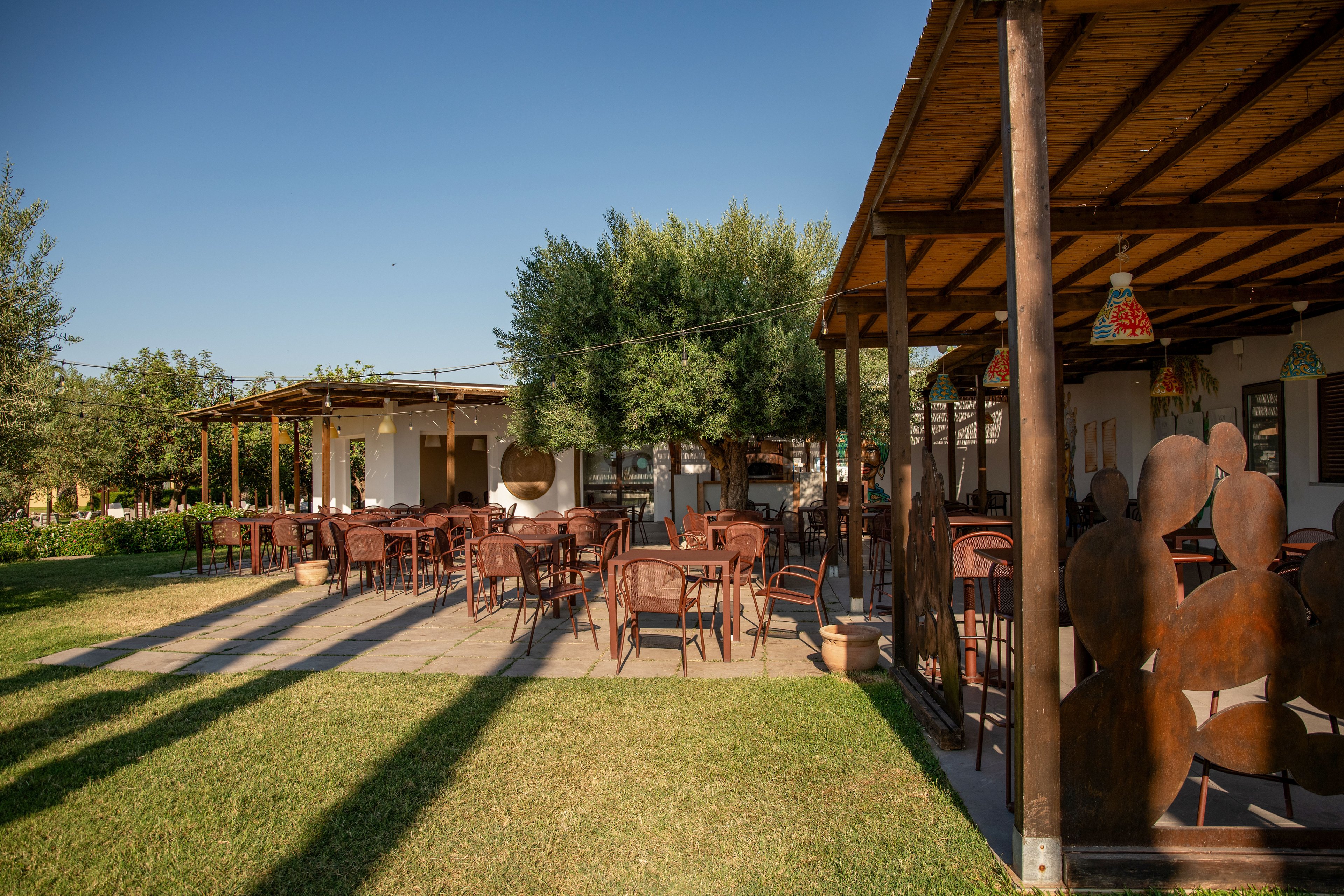 An open-air restaurant terrace features numerous wooden tables and chairs arranged on a paved patio. The space is covered by wooden pergolas with a thatched-style roof, providing shade and structure. Decorative elements and hanging lamps add character to the setting, while green grass and trees surround the dining area. The clear blue sky and warm sunlight create a relaxed, inviting atmosphere for outdoor dining.