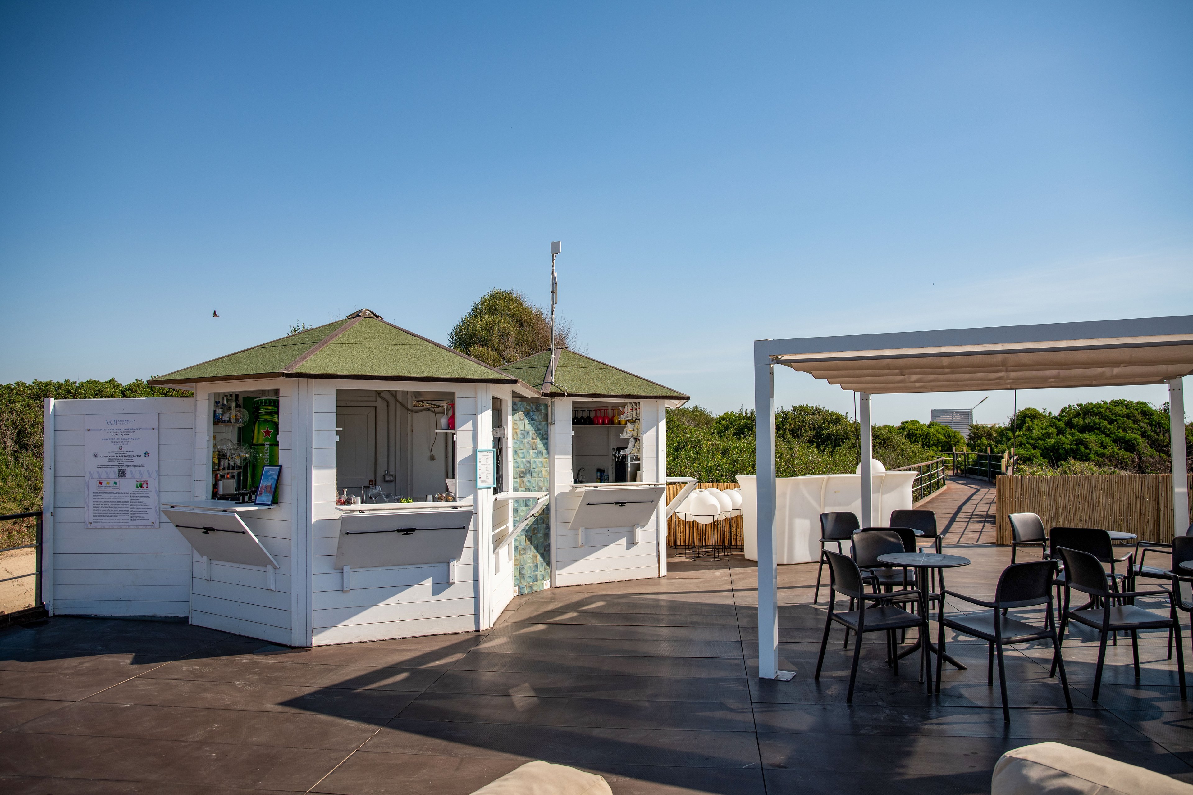 A small white kiosk stands on a spacious outdoor deck under a clear blue sky. The kiosk features open service windows and a shaded green roof, suggesting a casual refreshment stand. Nearby, a pergola covers several empty tables and chairs, creating a relaxed seating area. In the background, greenery and a sandy path lead toward the beach, evoking a calm coastal atmosphere.