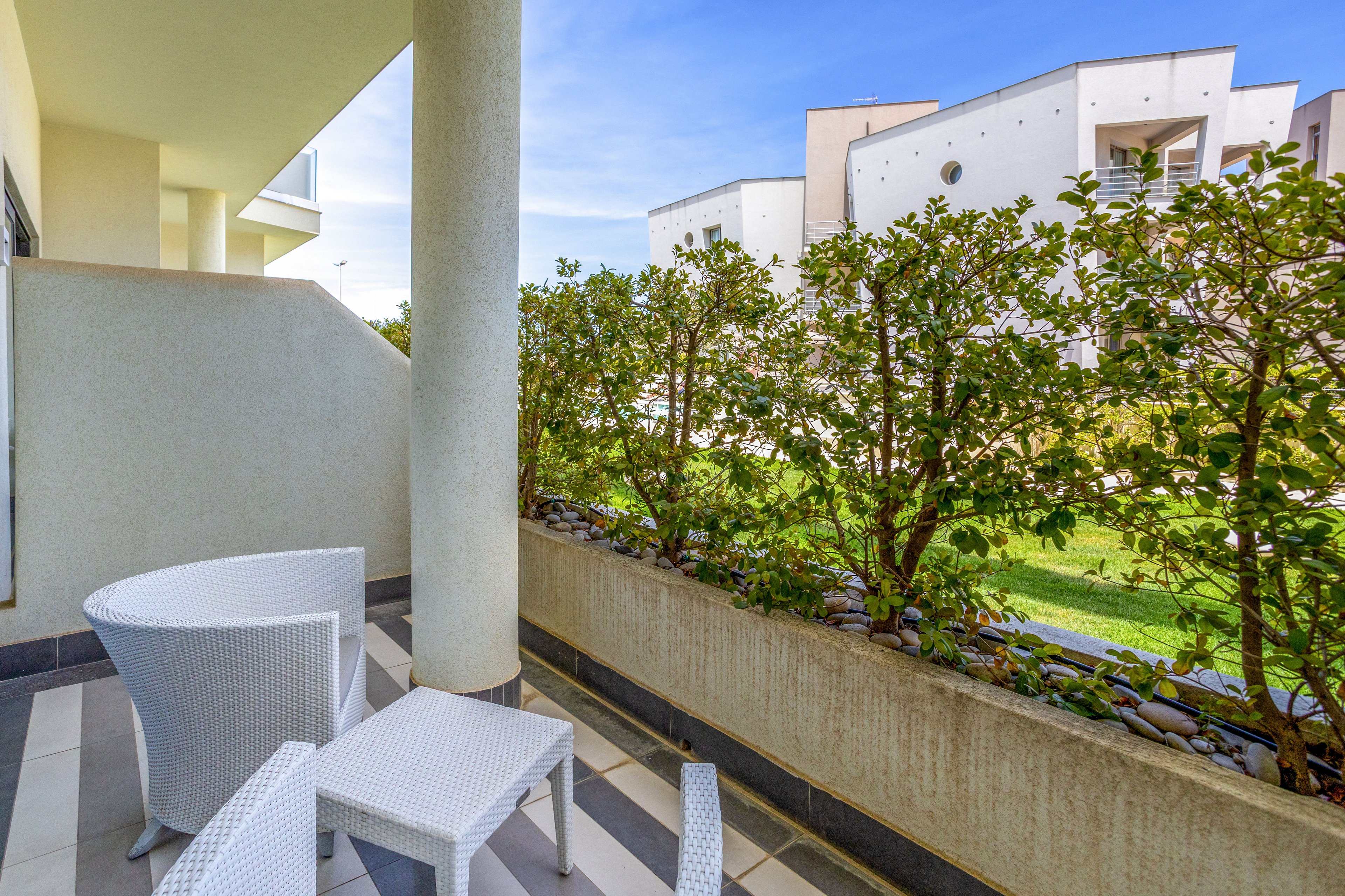 A contemporary apartment terrace features white outdoor chairs and a small table on a striped tiled floor. A tall column and beige walls frame the space, creating a clean, minimalist look. A neatly trimmed hedge lines the edge of the terrace, opening onto a green lawn and modern residential buildings in the background. The bright daylight and blue sky give the scene a fresh, inviting atmosphere.