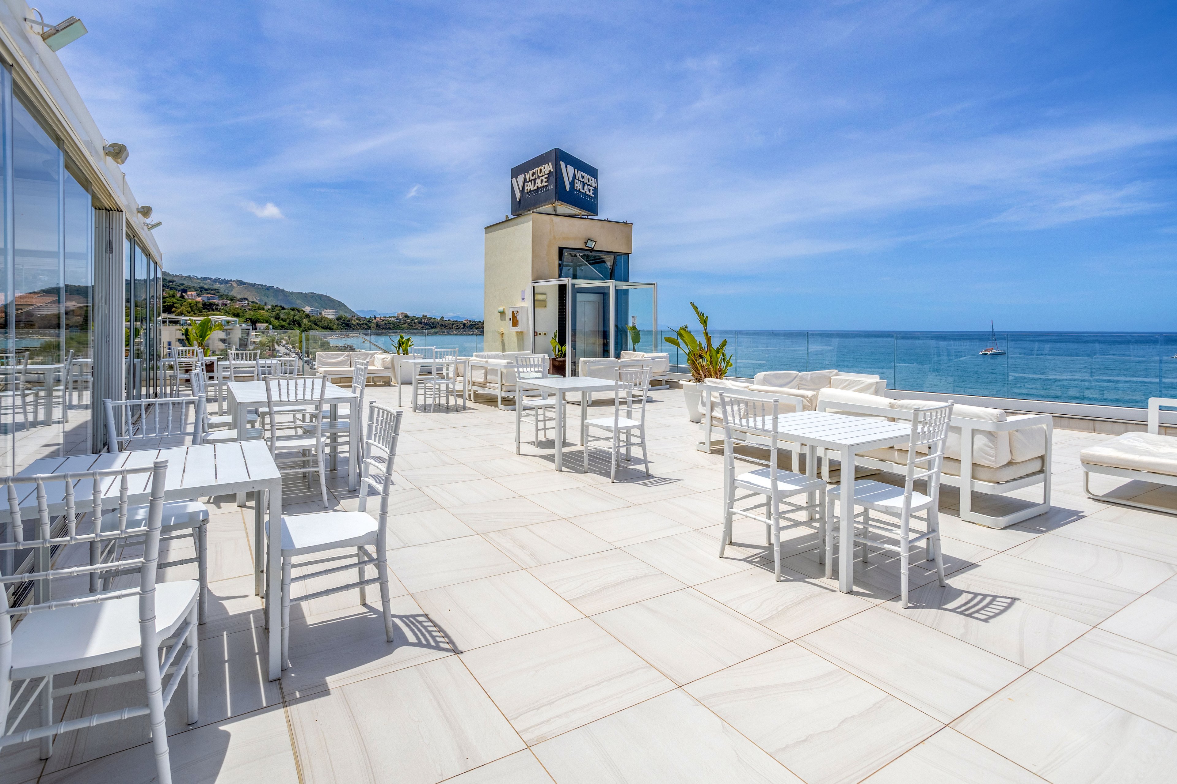 An open-air rooftop terrace overlooks a calm blue sea under a bright, clear sky. White tables, chairs, and cushioned sofas are arranged across a spacious tiled floor, creating a clean and modern look. Glass railings and a mirrored wall reflect the coastal landscape and nearby hillside buildings. A small tower structure with visible text stands near the center, adding architectural interest to the relaxed waterfront setting.