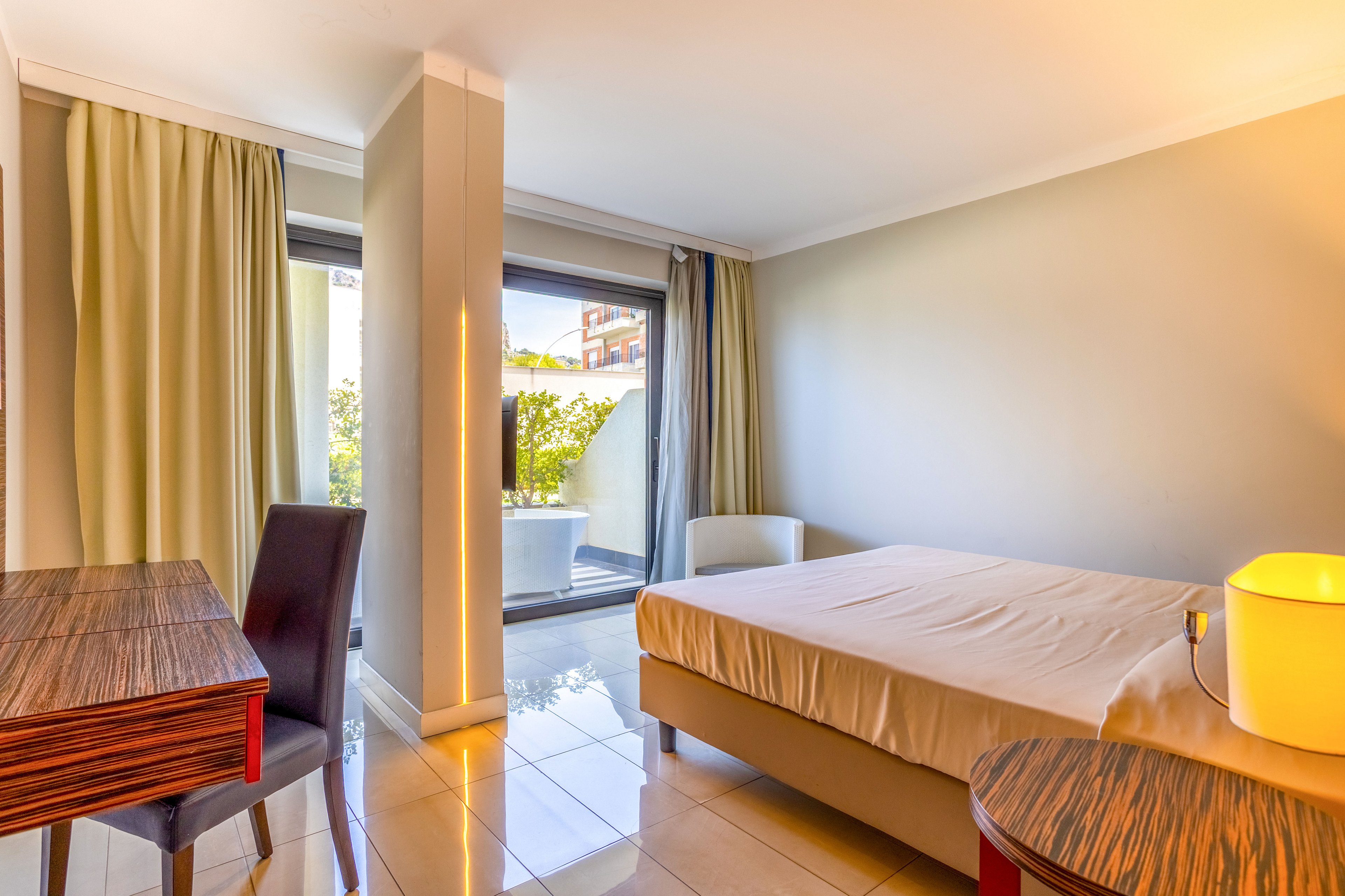 A contemporary hotel room features a neatly made bed and glossy tiled floor. Large sliding glass doors open to a small terrace with greenery and outdoor light. A wooden desk and round side table add warm tones, while neutral walls and curtains create a calm atmosphere. A modern lamp on the side table provides additional soft lighting.