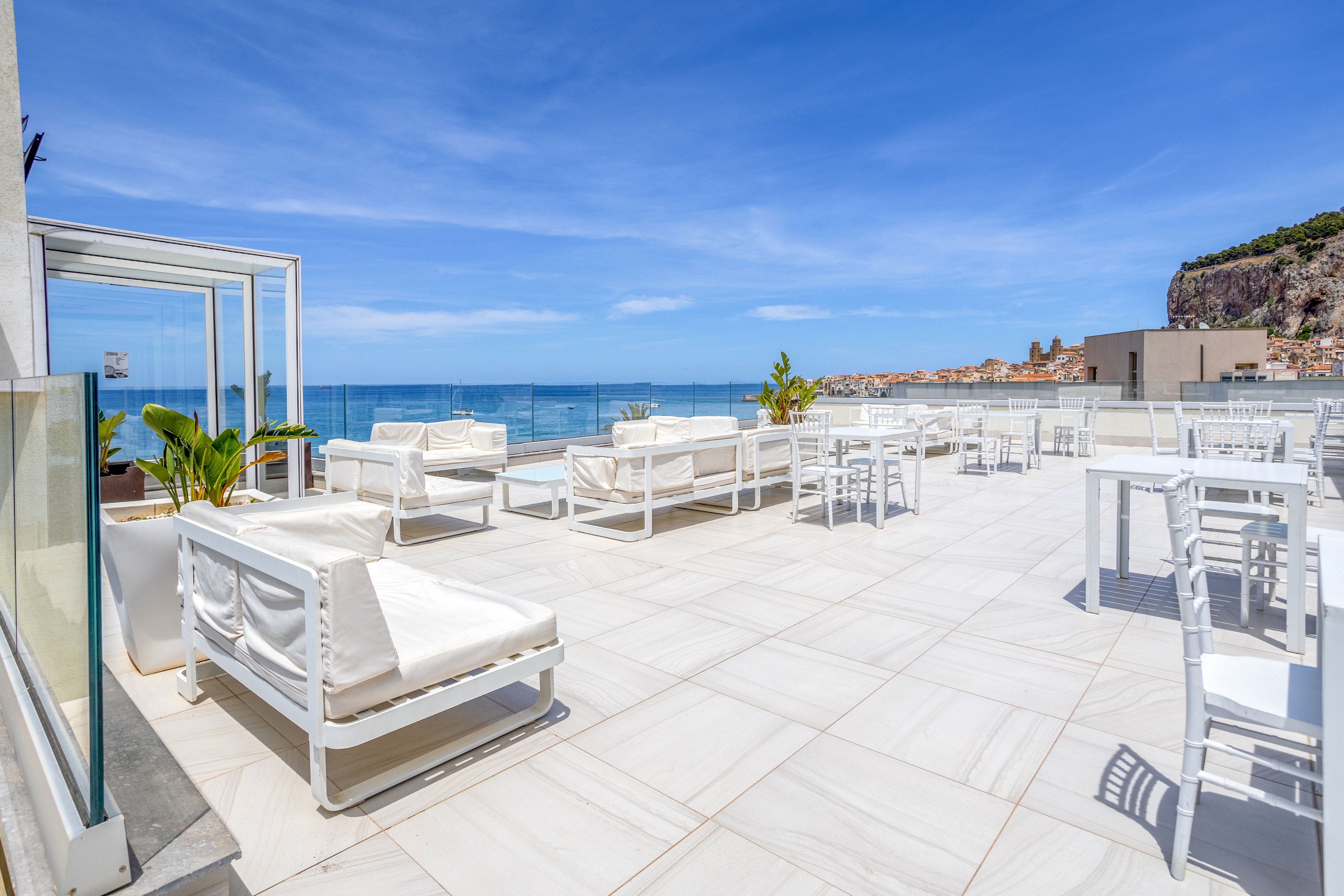 A bright rooftop terrace features white lounge sofas and dining tables under a clear blue sky. The space is open and spacious, with tiled flooring and glass railings framing the view. In the background, the calm sea and coastal town are visible along the shoreline. Potted plants add a touch of greenery to the otherwise minimalist, modern setting.
