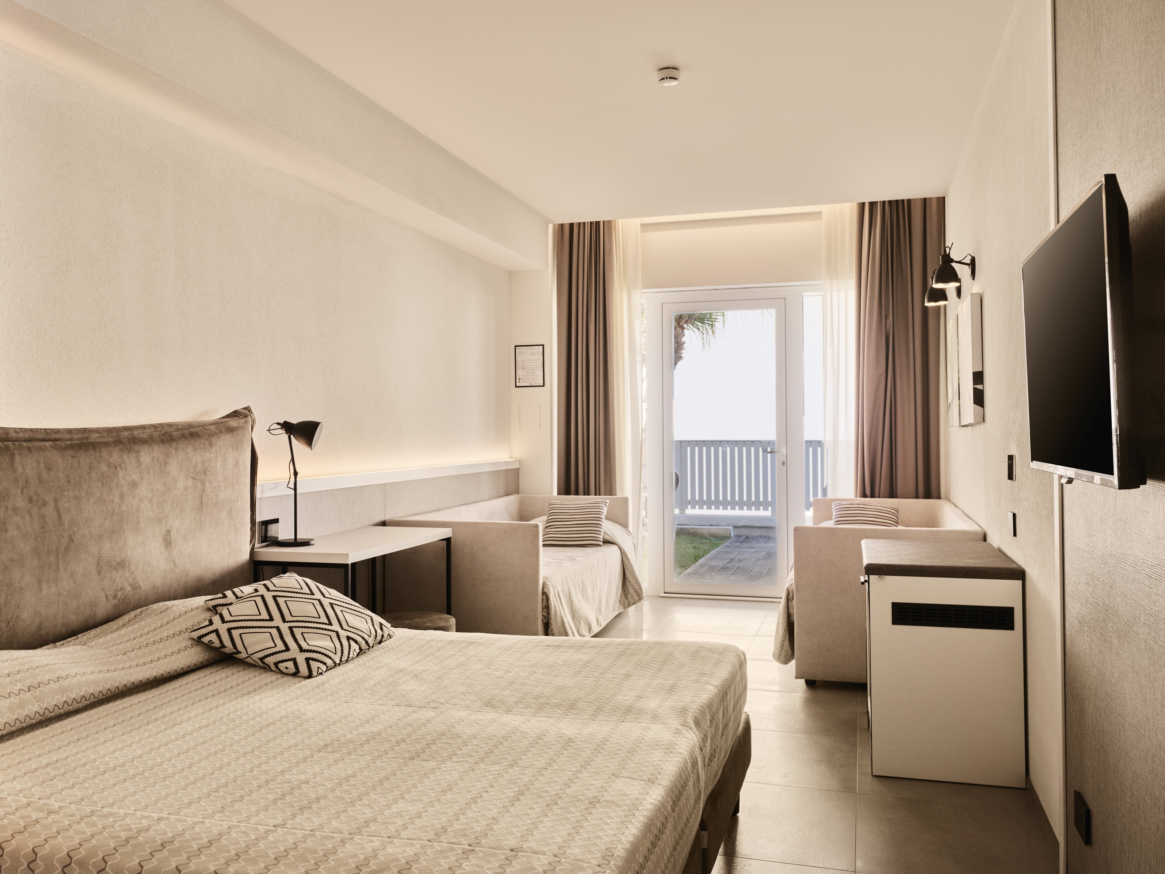 A contemporary hotel room features a large bed in the foreground with a patterned cushion and neutral bedding. A desk with a reading lamp and an additional single bed sit along the left wall, while a wall-mounted television and compact cabinet are on the right. Floor-to-ceiling curtains frame glass doors that open to a balcony with a railing and a glimpse of greenery outside. The space is decorated in soft beige and gray tones, creating a clean, minimalist and inviting atmosphere.