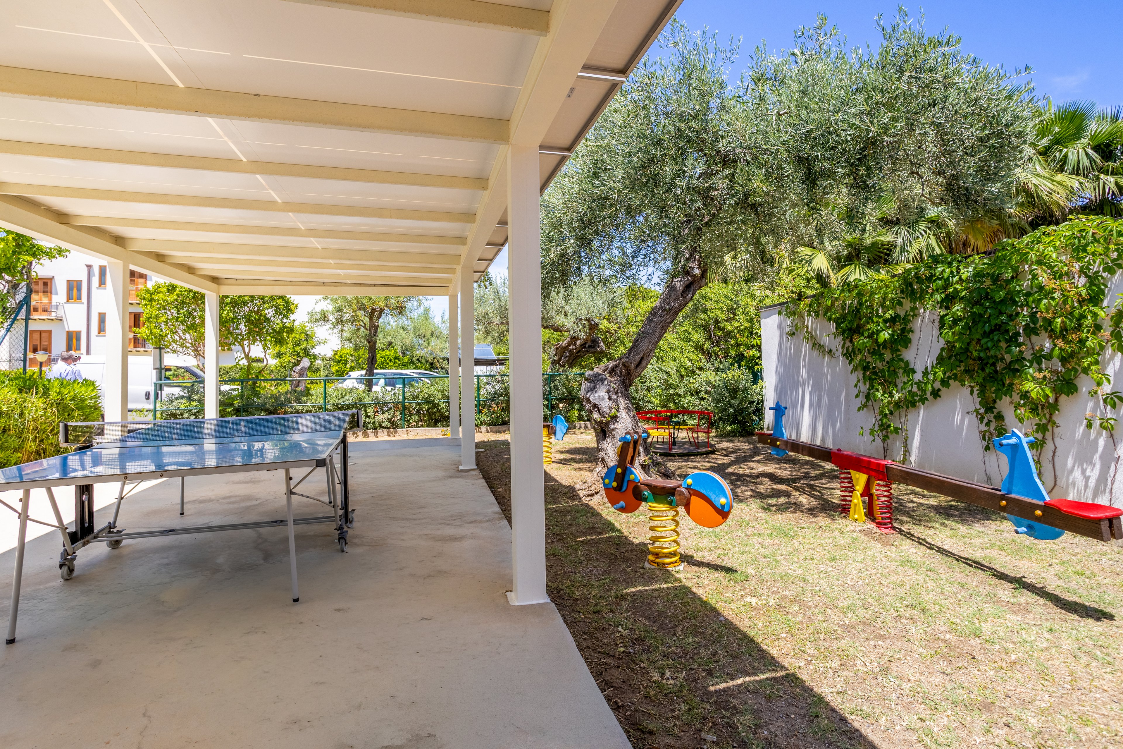 An outdoor recreation area features a covered ping pong table beside a small playground. Colorful spring-mounted rocking toys and seesaws sit on a grassy patch under a large tree. A white fence with lush green vines borders the space, with residential buildings visible in the background. The scene is bright and sunny, suggesting a family-friendly leisure environment.