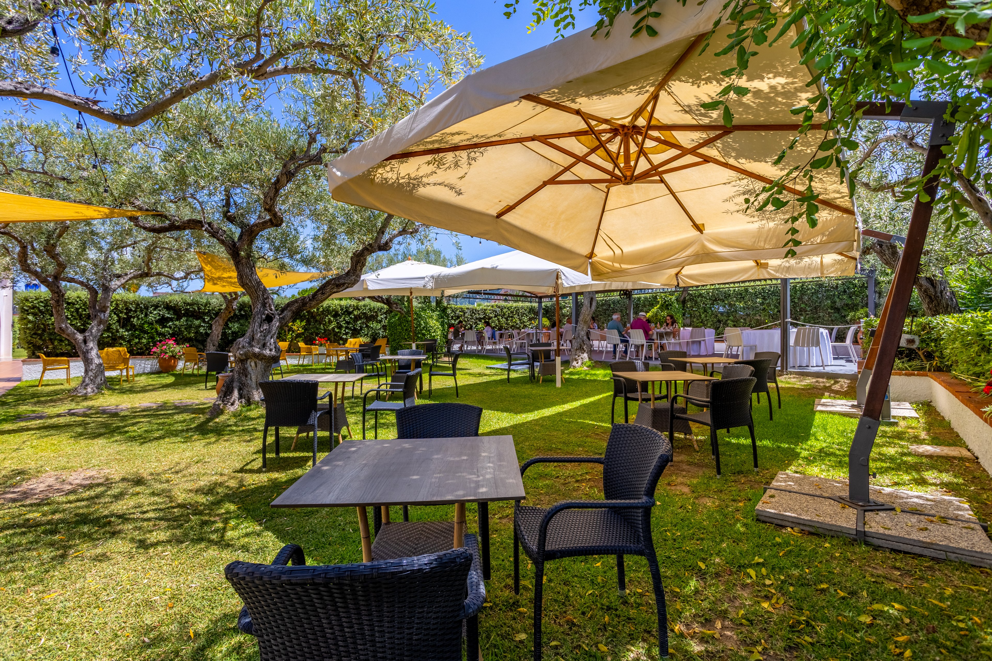 An inviting outdoor restaurant terrace is set on a grassy lawn beneath large shade umbrellas. Black wicker chairs and dark tables are arranged across the garden, with more seating visible in the background. Mature trees, hedges, and flowering plants surround the space, creating a relaxed and natural atmosphere. A few people can be seen near the far tables, suggesting a casual dining or social gathering setting.
