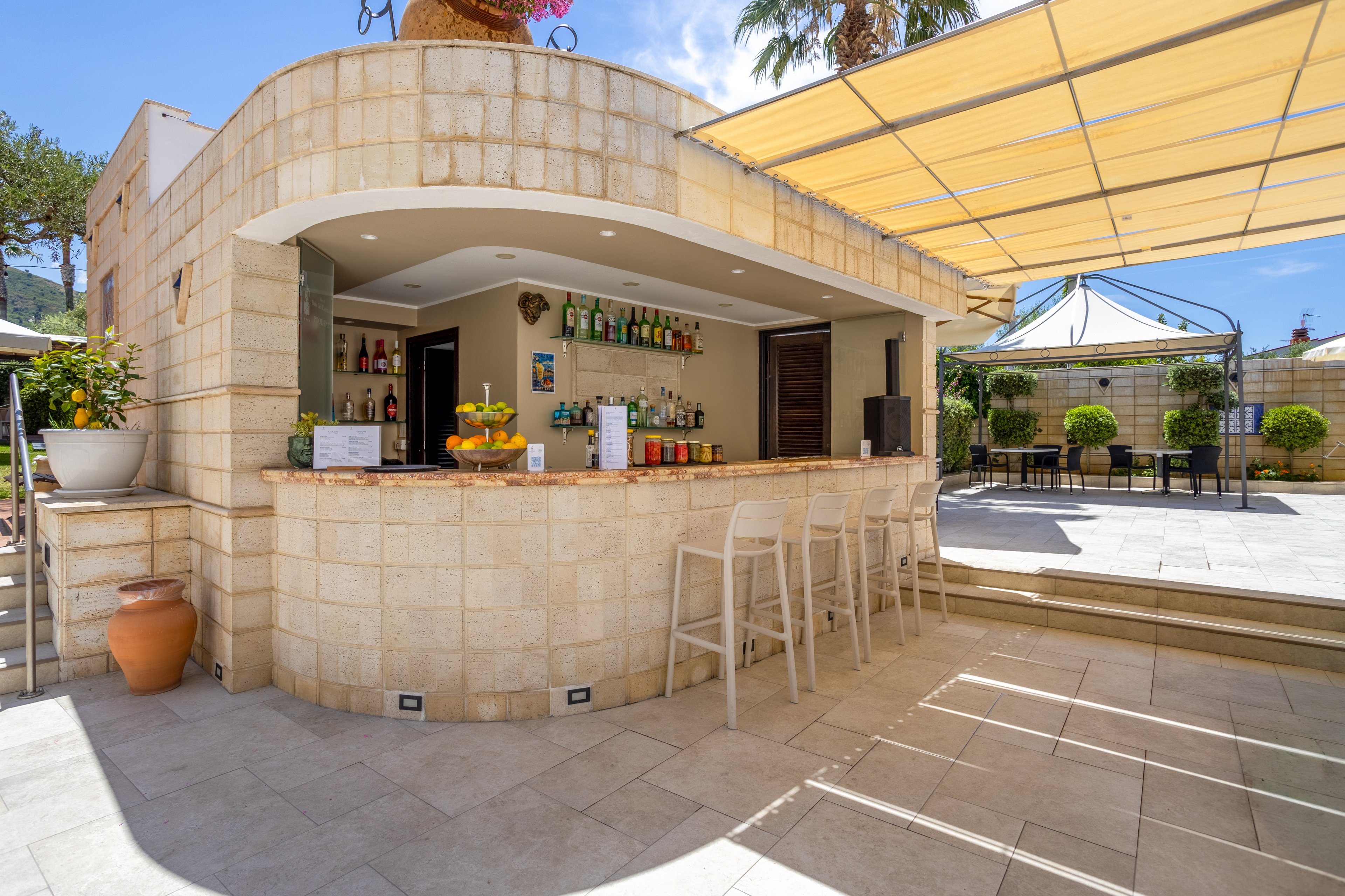 An open-air bar with a stone counter stands in a sunny courtyard, stocked with colorful bottles and glassware. Several transparent bar stools line the counter, while a fruit bowl adds a fresh accent to the scene. A large canopy provides shade over the tiled terrace, with tables and chairs arranged for guests. The setting suggests a relaxed vacation atmosphere in a warm, bright climate.