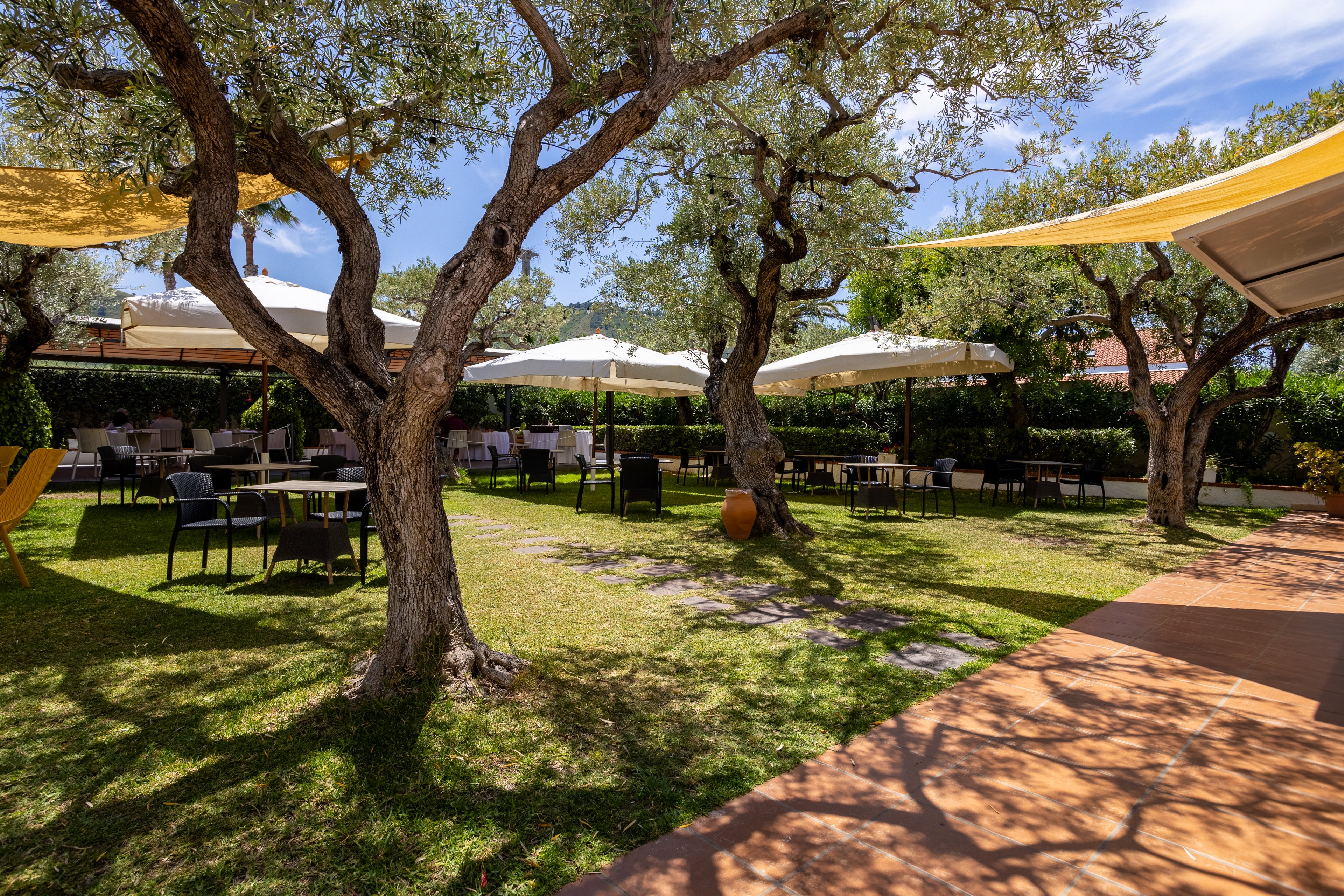 An inviting outdoor dining area is set in a lush garden with mature olive trees casting dappled shade. Tables and chairs are arranged across the grass beneath large white umbrellas. Yellow fabric awnings add warm color and additional shade along the patio edge. The scene suggests a relaxed, Mediterranean-style atmosphere on a bright, sunny day.
