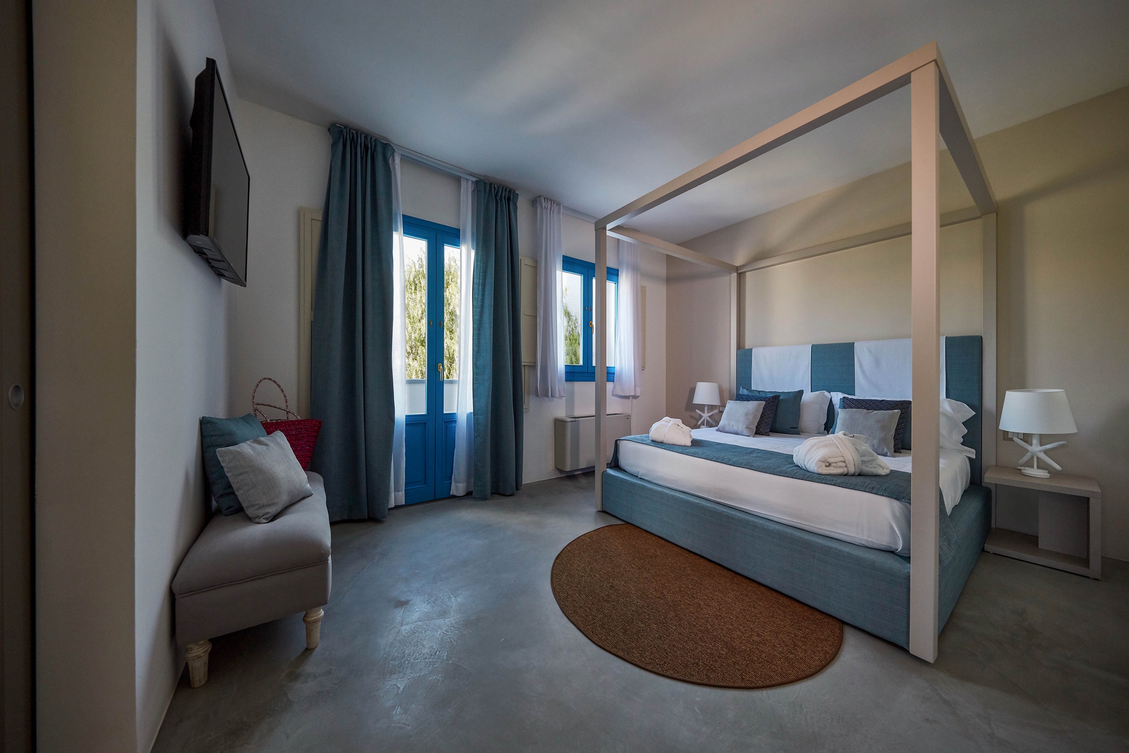 A contemporary bedroom features a large canopy bed with blue and white bedding as the focal point. The room has concrete floors, blue curtains, and tall windows letting in natural light. A small upholstered bench with cushions sits beneath a wall-mounted television. A bedside table with a white lamp and a round brown rug add warmth to the minimalist decor.