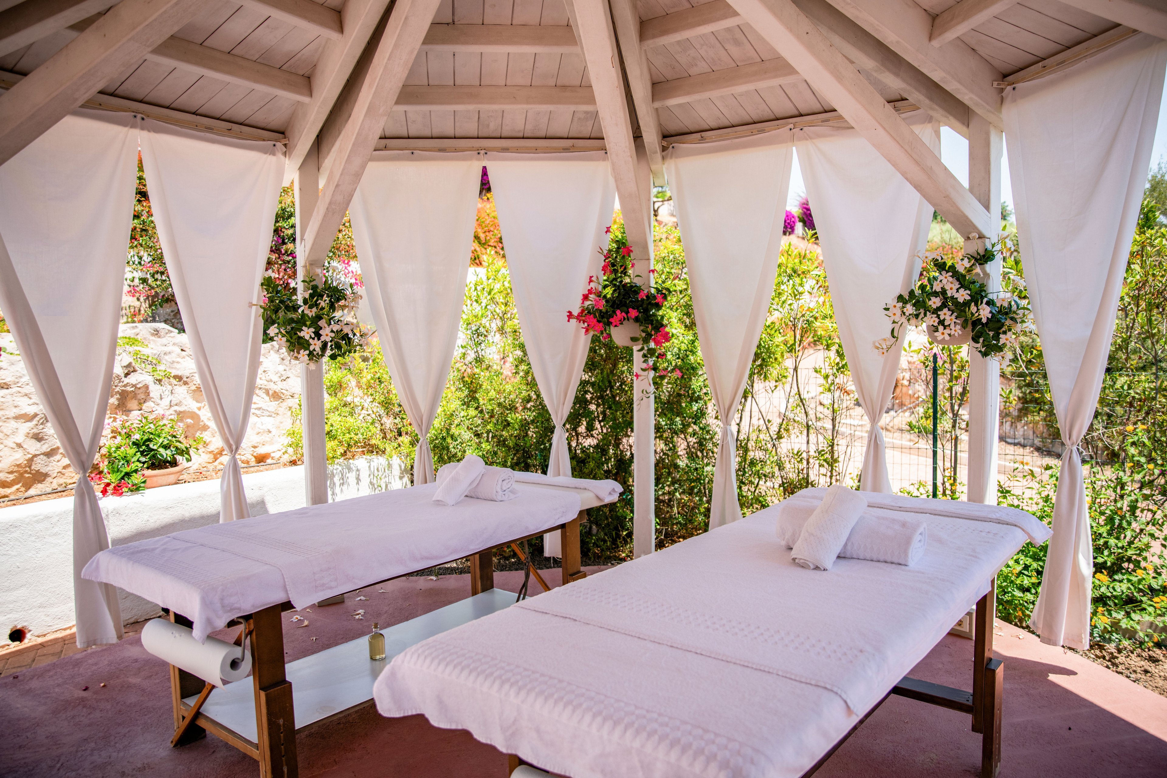 Two massage tables covered in white linens sit inside an open-air cabana. White curtains hang between wooden beams, creating a private and serene spa setting. Colorful flowers decorate the structure, and greenery is visible in the background. The bright daylight and soft pastel tones convey a relaxing, luxurious atmosphere.